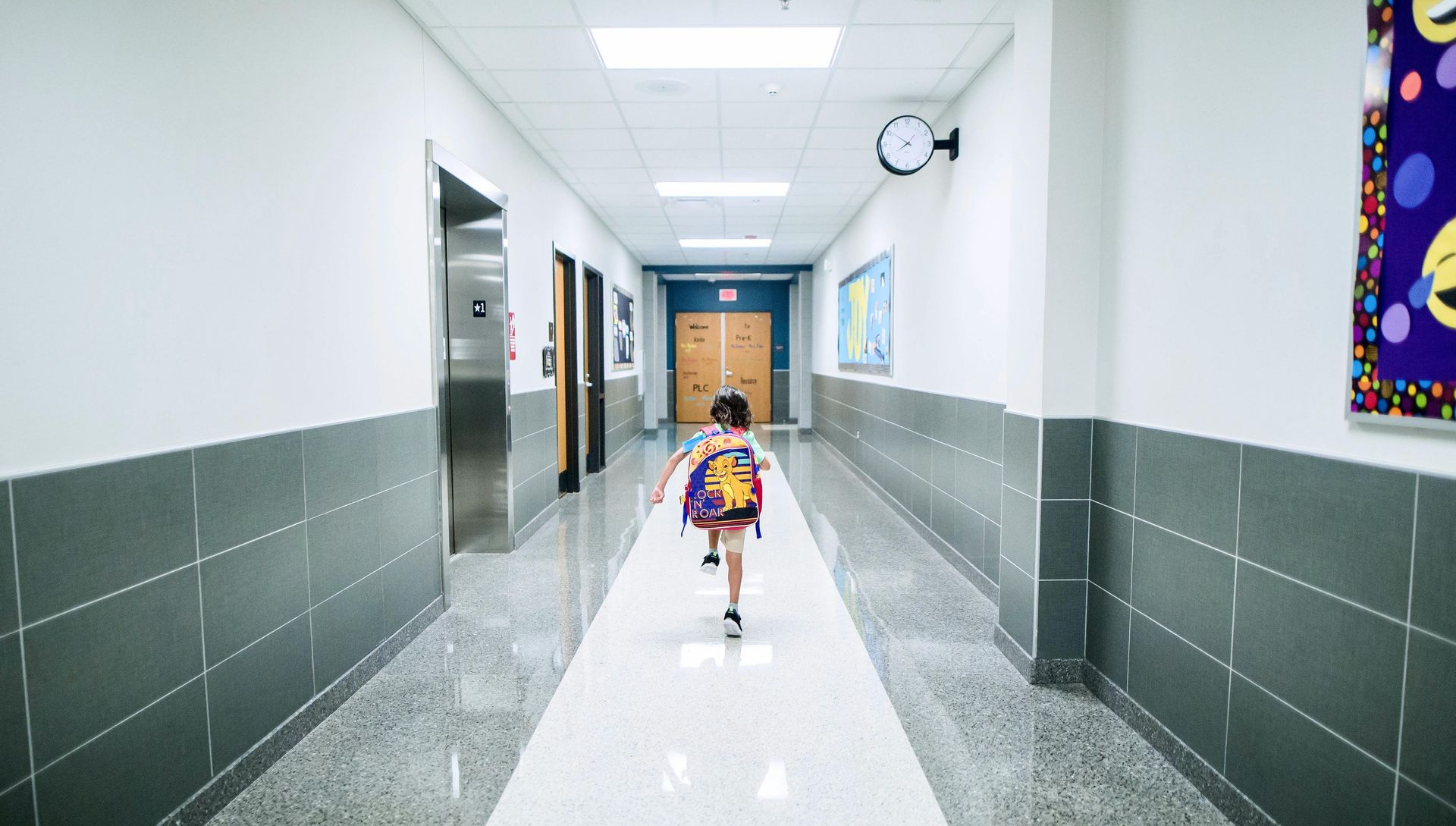 Child running down a long school hallway, carrying a backpack.