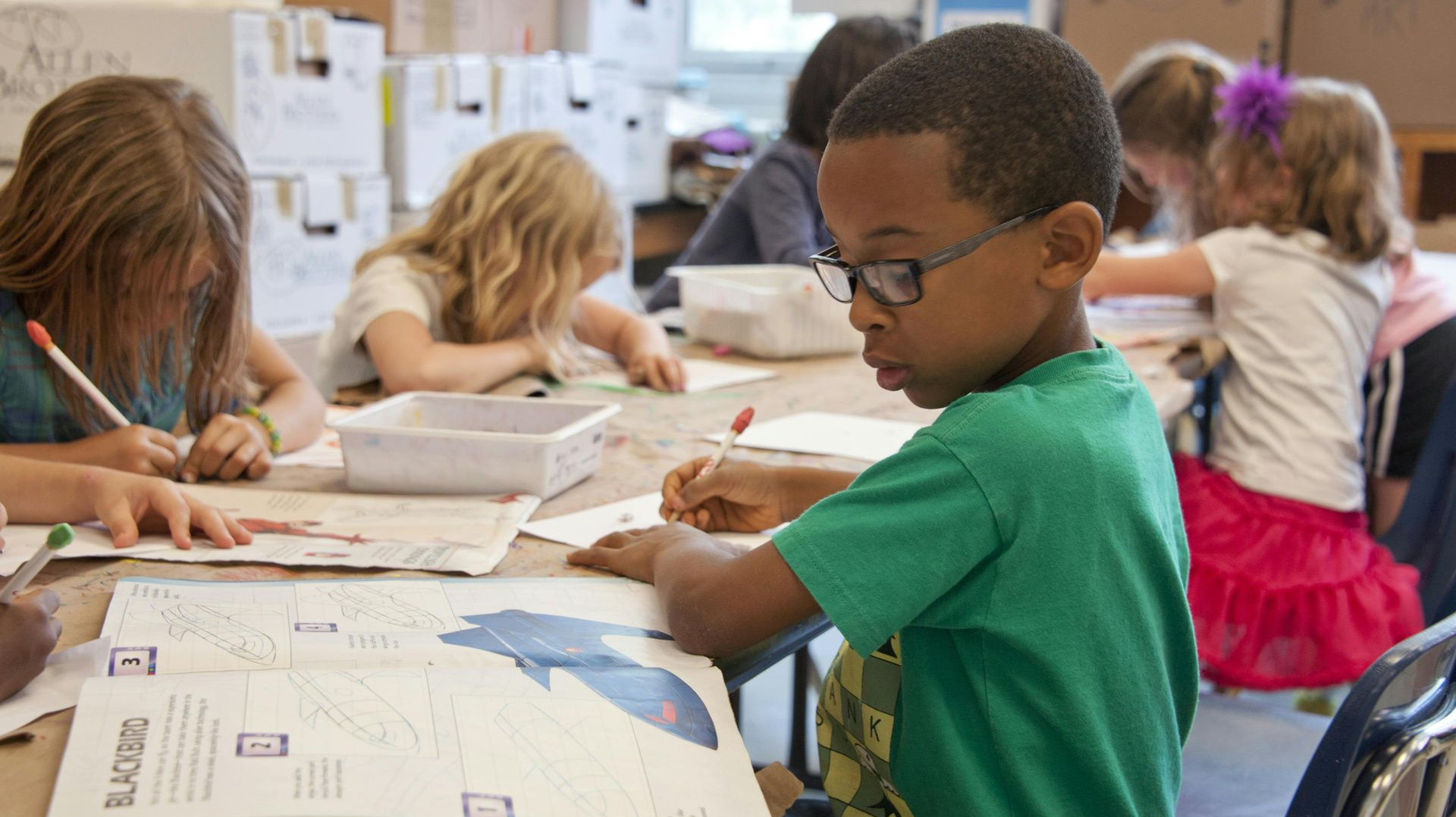 Children in a classroom, focused on work at a table. One boy in a green shirt and glasses writes in a workbook.