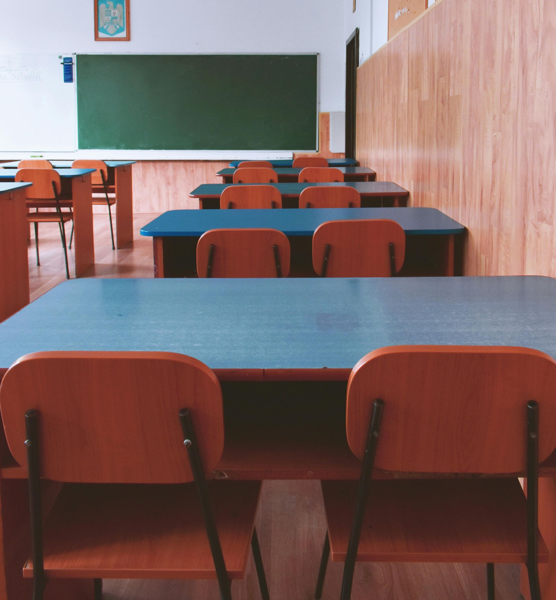 Empty classroom with rows of desks, chairs, and a chalkboard. Wooden walls, blue tabletops, and orange chairs.