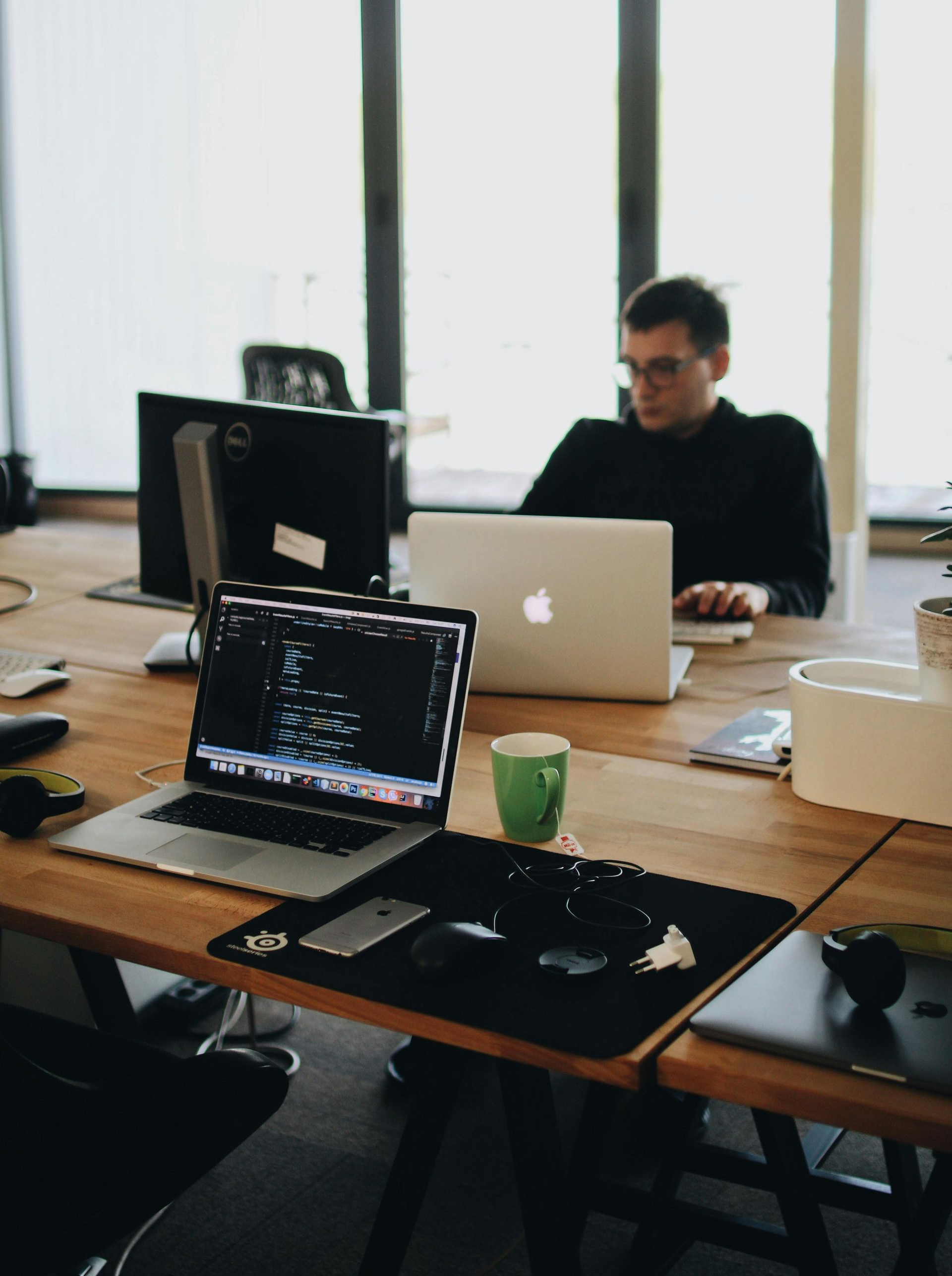 A person working on a laptop at a shared wooden desk, other laptops and office supplies visible.