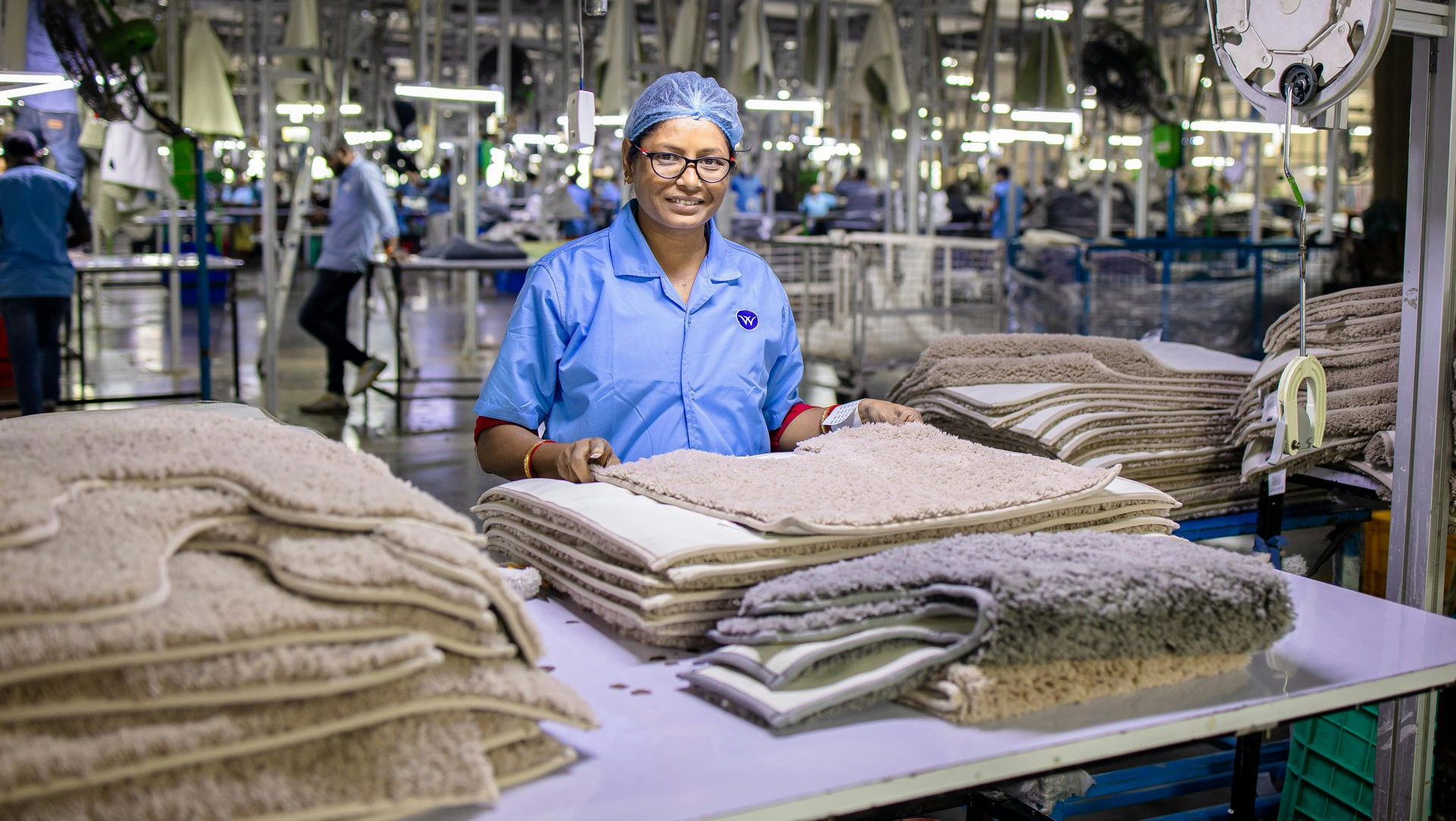 Woman in blue uniform smiles at camera, standing amid stacks of beige and gray rugs in a factory.