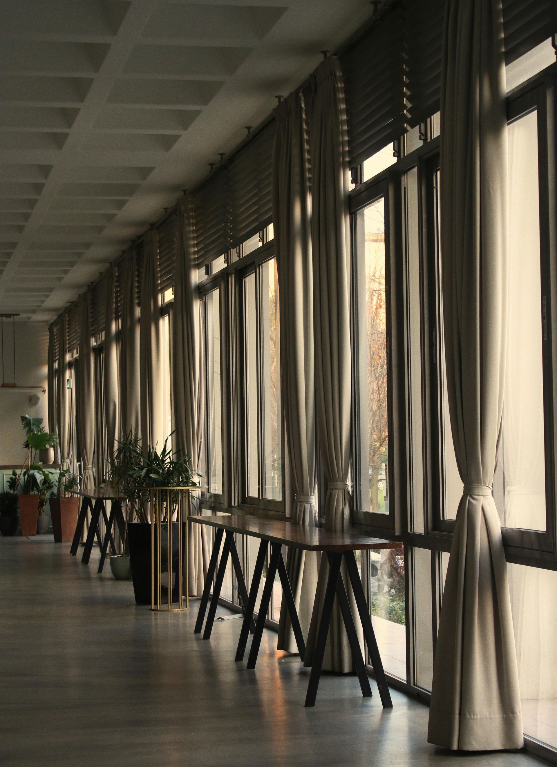 Long hallway with floor-to-ceiling windows draped with tan curtains. Tables are set up along the windows.