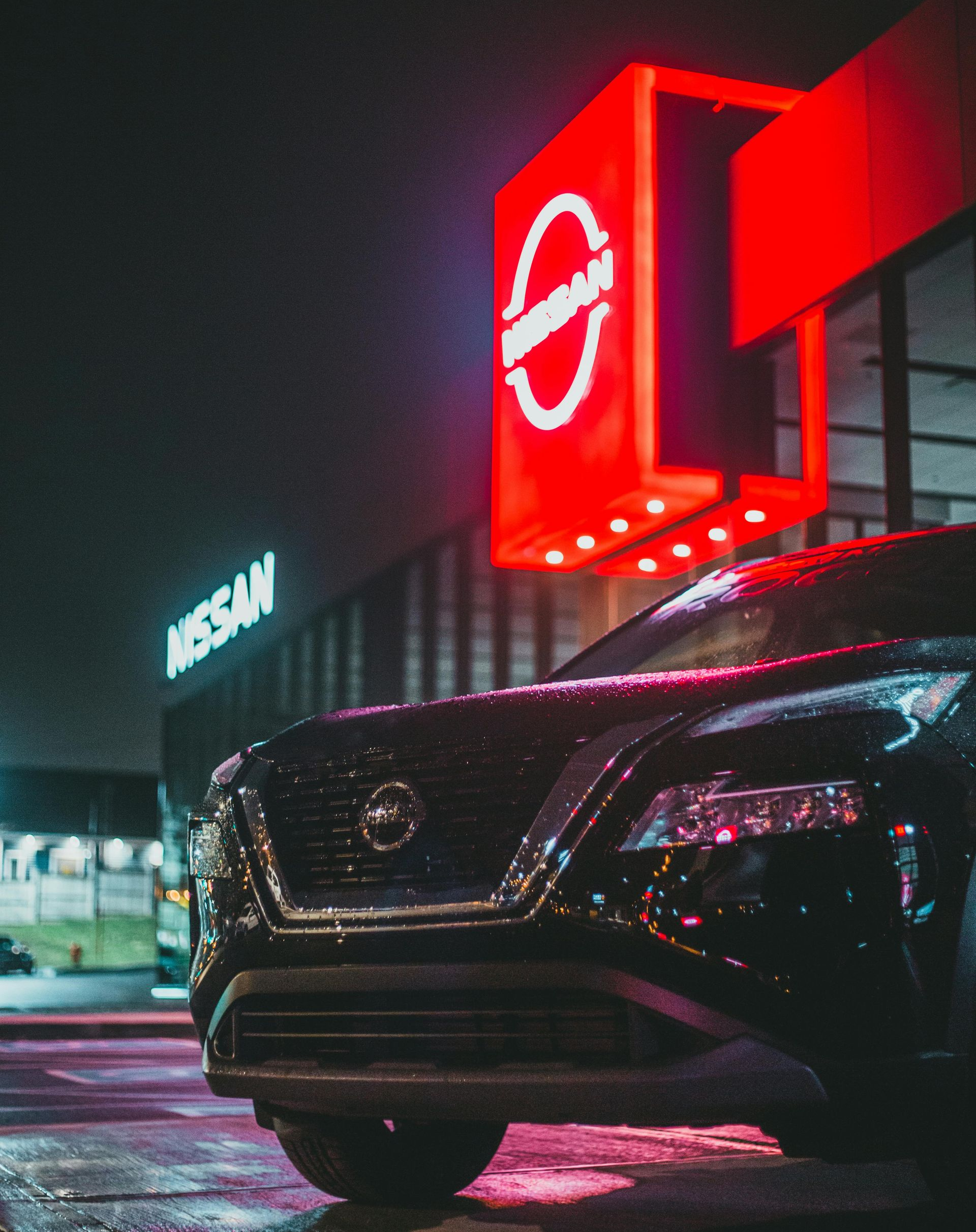 Black Nissan SUV parked in front of a neon red Nissan dealership sign at night.