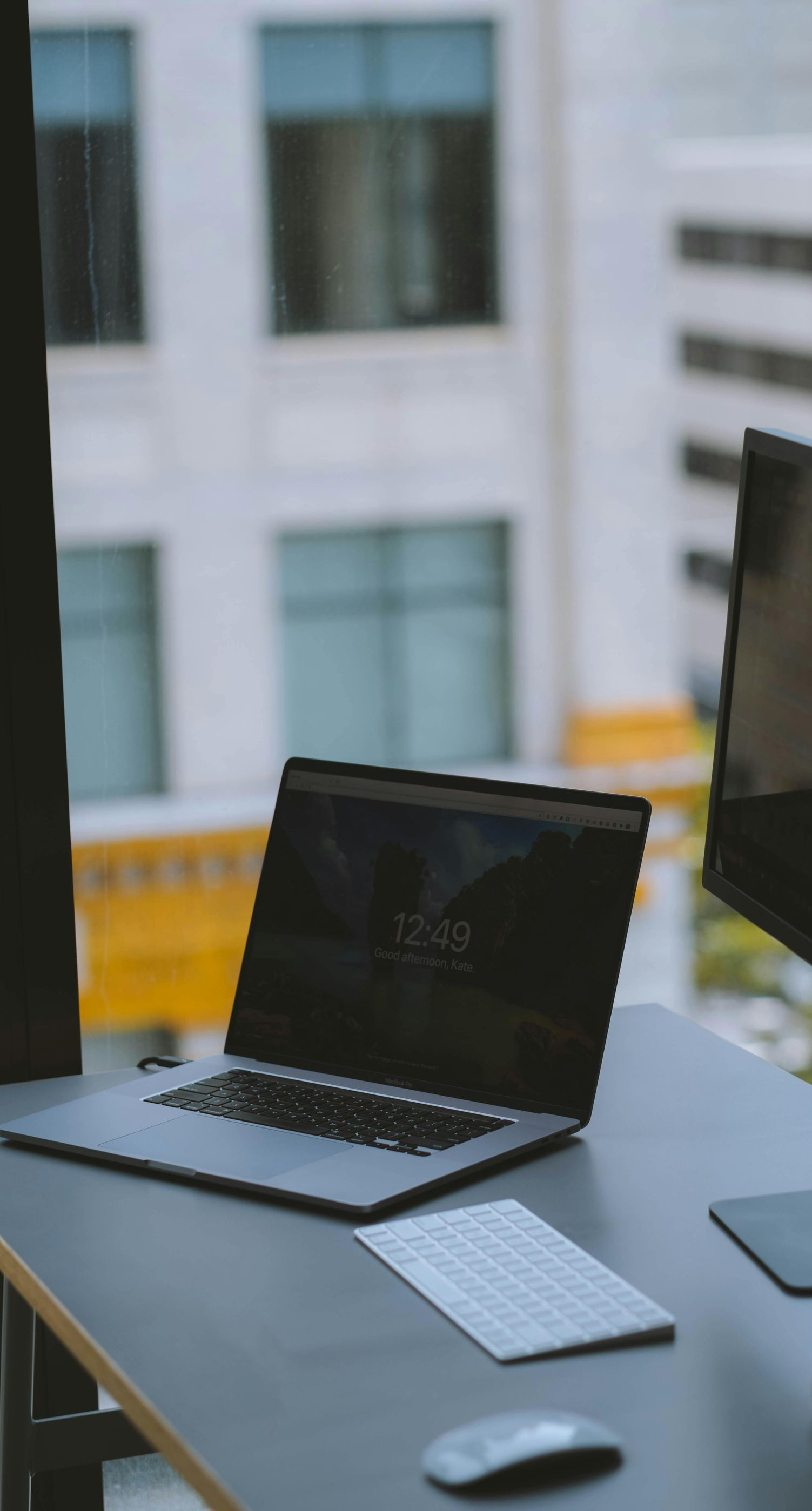 Laptop, keyboard, and mouse on a desk by a window; a second monitor and coffee cup visible.