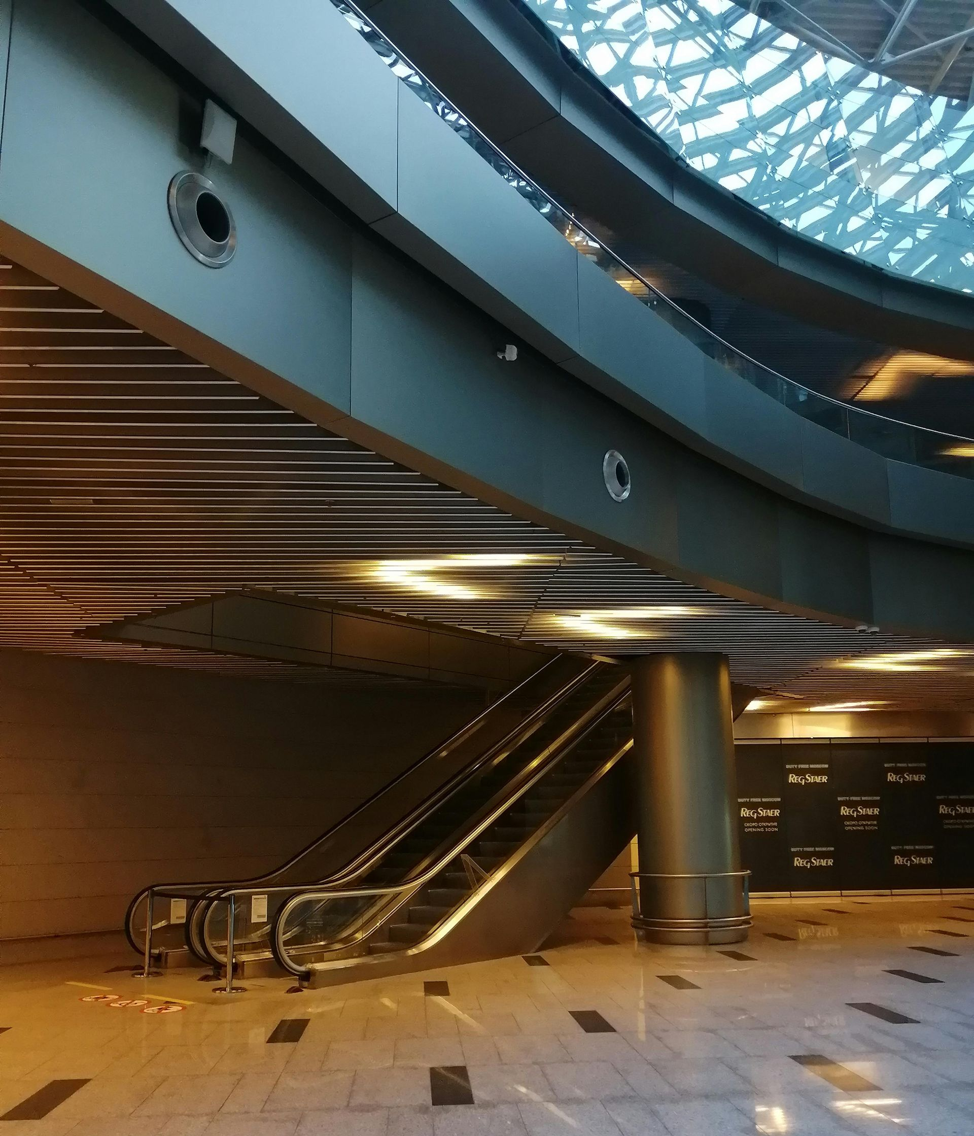 Escalator in a modern building with curved ceiling, glass roof, and ambient lighting.