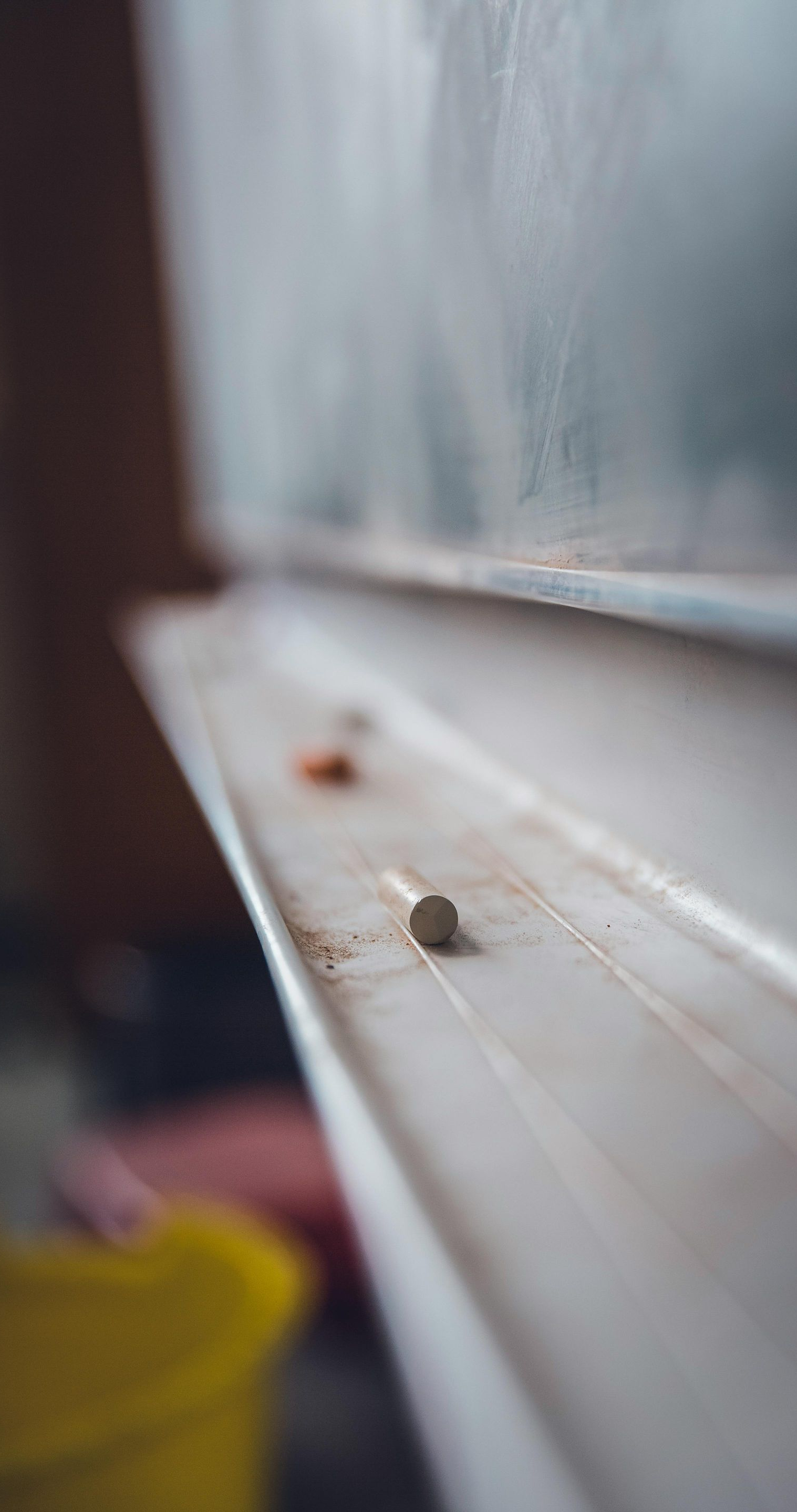 Blackboard with chalk in a tray. Focus on chalk and the blackboard’s edge. Yellow bucket visible in the blurred foreground.