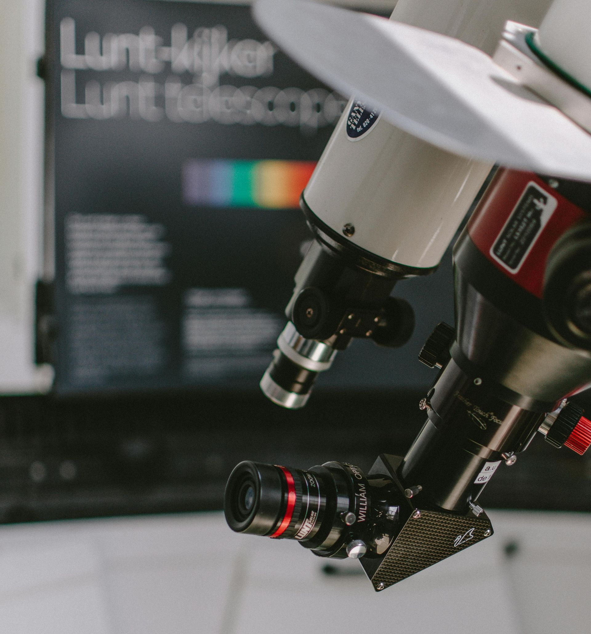 Telescopes with attached camera equipment, near a color spectrum chart and black table, in a bright room.