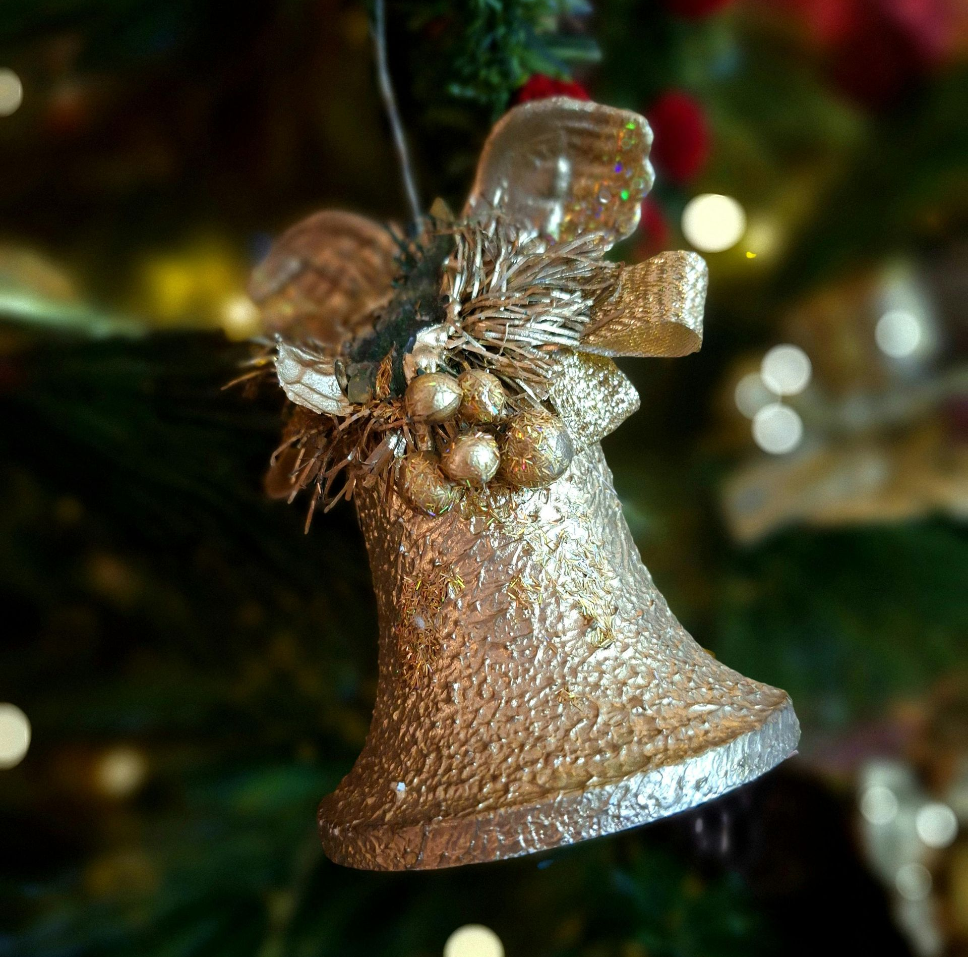 Gold Christmas bell ornament with textured surface, bow, and berries, hanging on a tree.