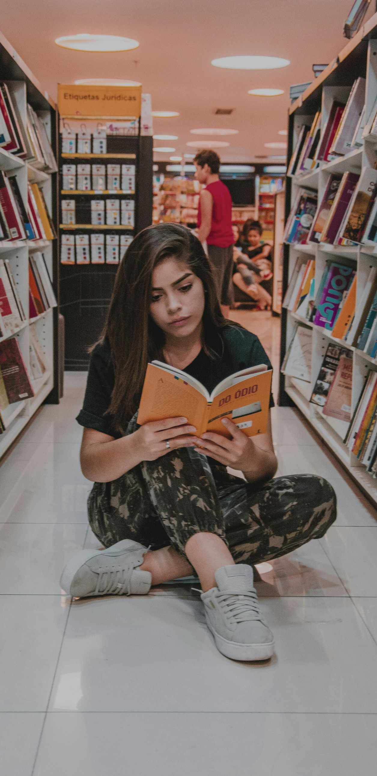 Woman reading a book while sitting on the floor in a library, surrounded by bookshelves.