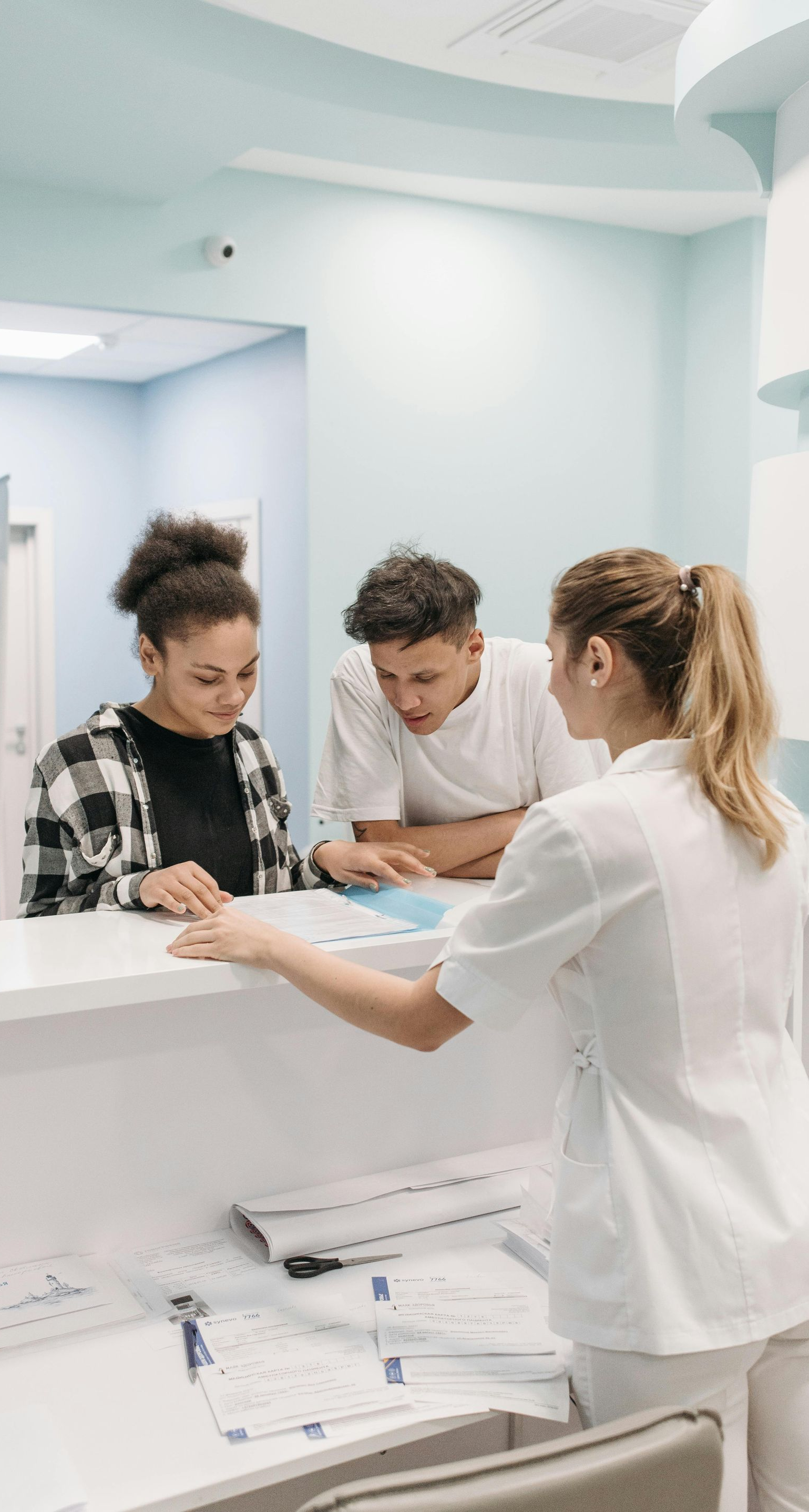 Two people at a reception desk, being assisted by a person in a white coat. Light blue walls, forms.