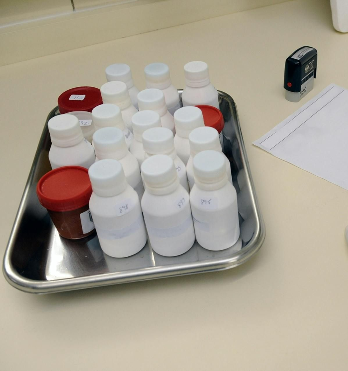 White pill bottles and two red-capped bottles sit in a metal tray; stamp and paper in the background.