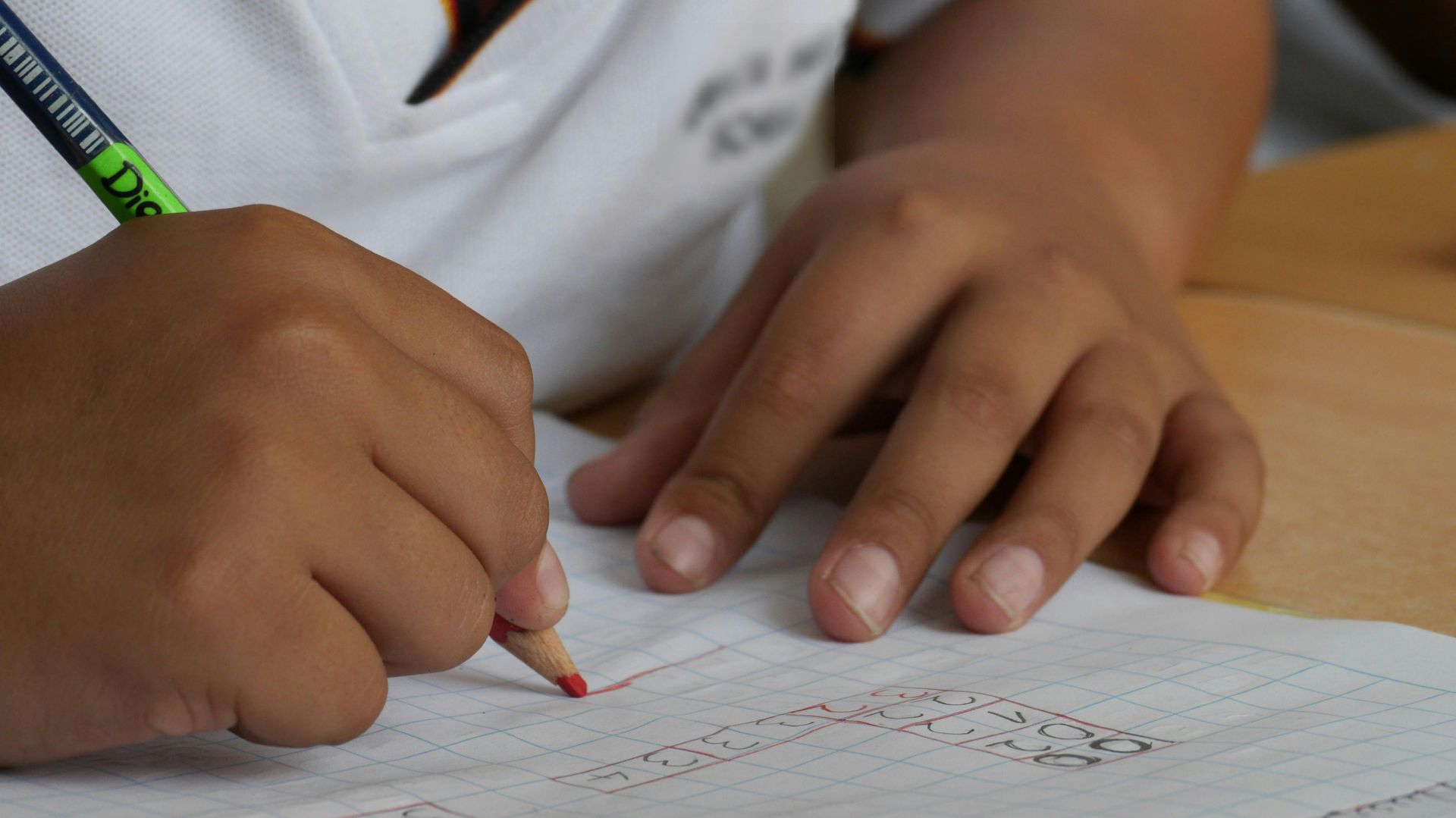A child's hands writing with a red pencil on graph paper, close-up.