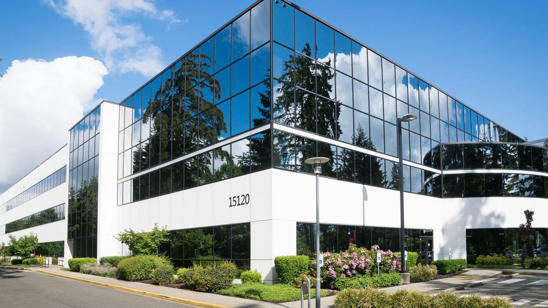 Modern white office building with large glass windows, trees reflected, and blue sky.