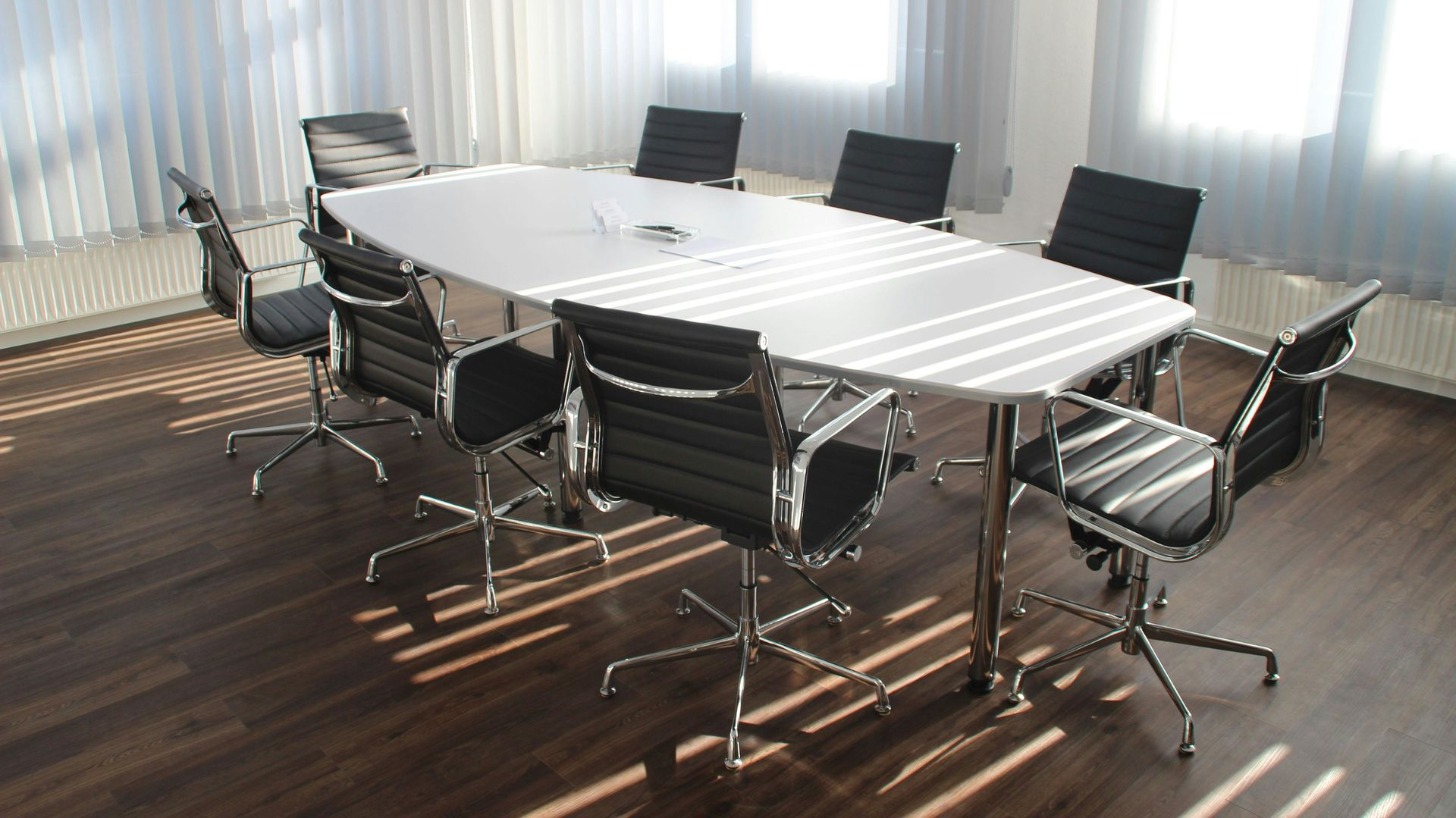 Conference room with oval white table and black chairs, sunlight through blinds.
