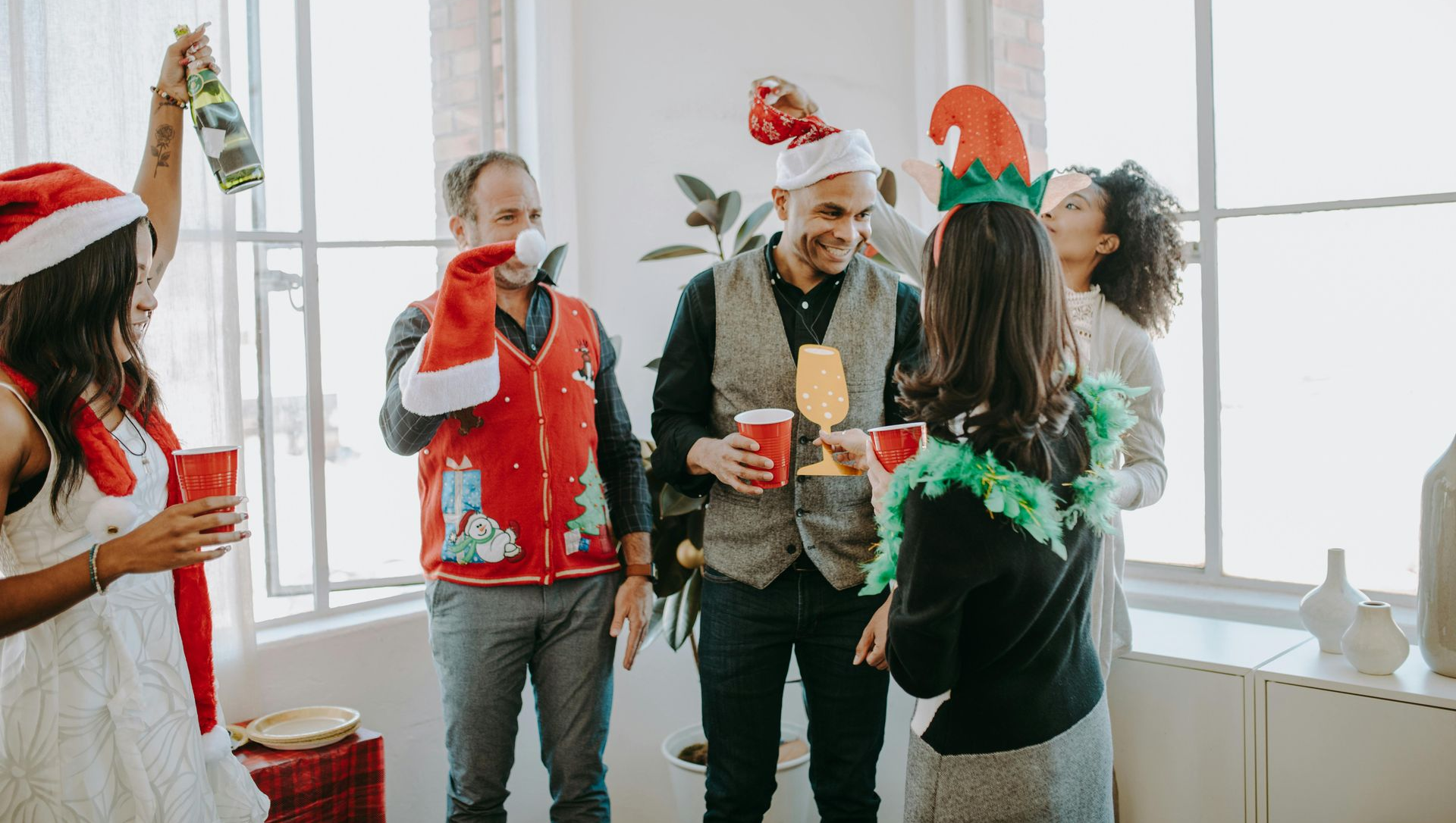 People in Christmas attire at a party, toasting with drinks. Room with windows.