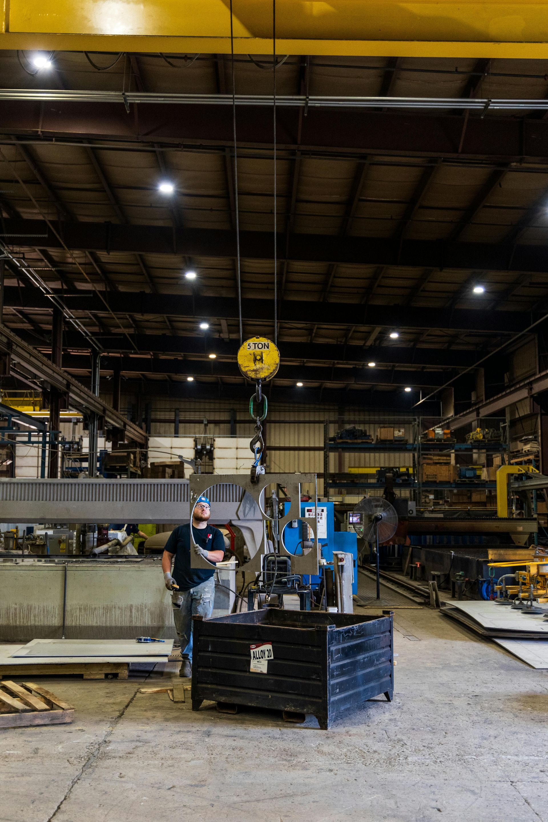 A person in a warehouse operates an overhead crane to lift a metal component into a black industrial bin.