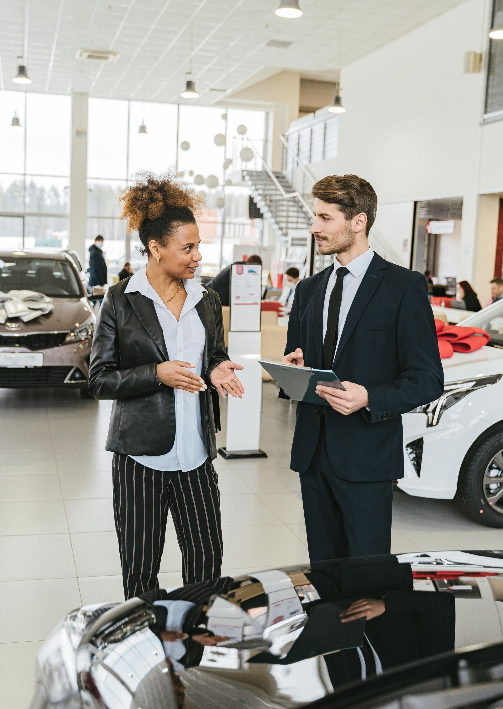 Woman in car dealership talking to salesperson, standing near a black car.