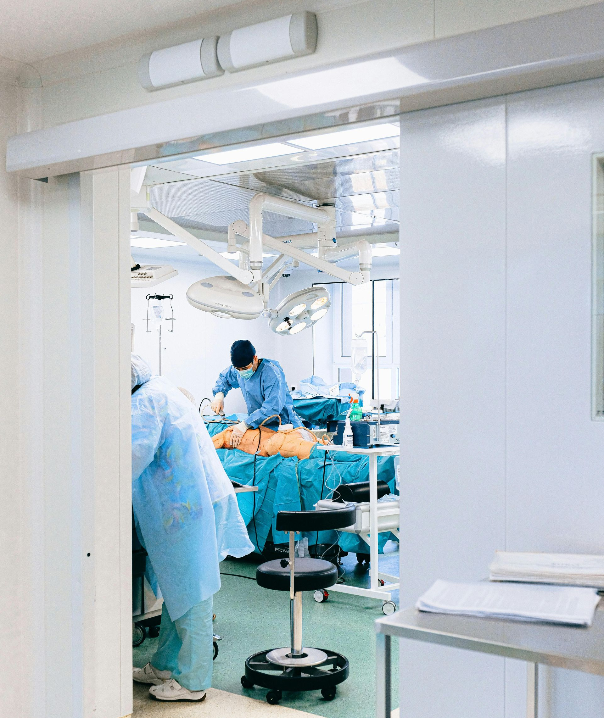 Operating room with surgeons performing surgery, viewed through doorway. Bright lighting, sterile environment.