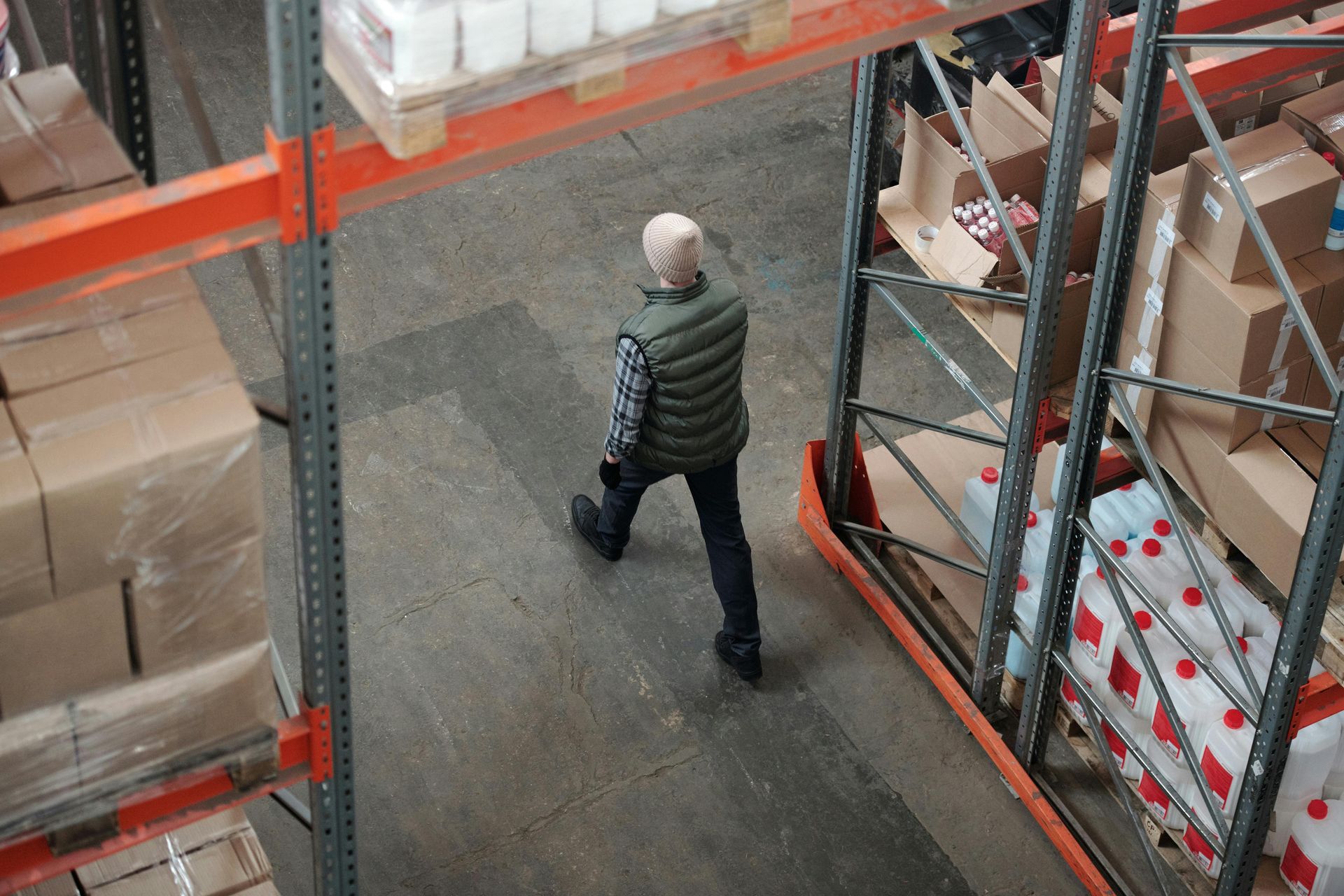 Person walking in a warehouse aisle with tall shelves of boxes. They wear a vest and dark pants.