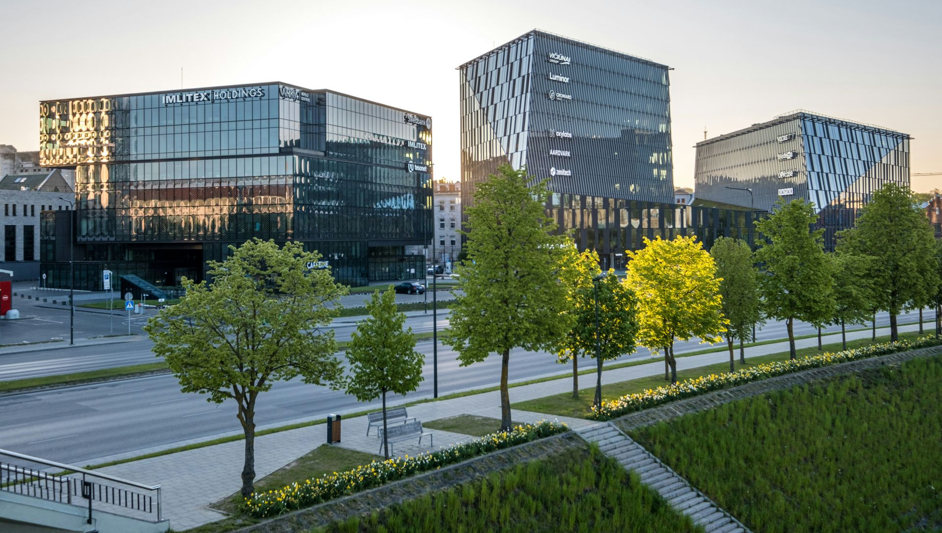 Modern glass buildings with trees and a road in front; sunset.