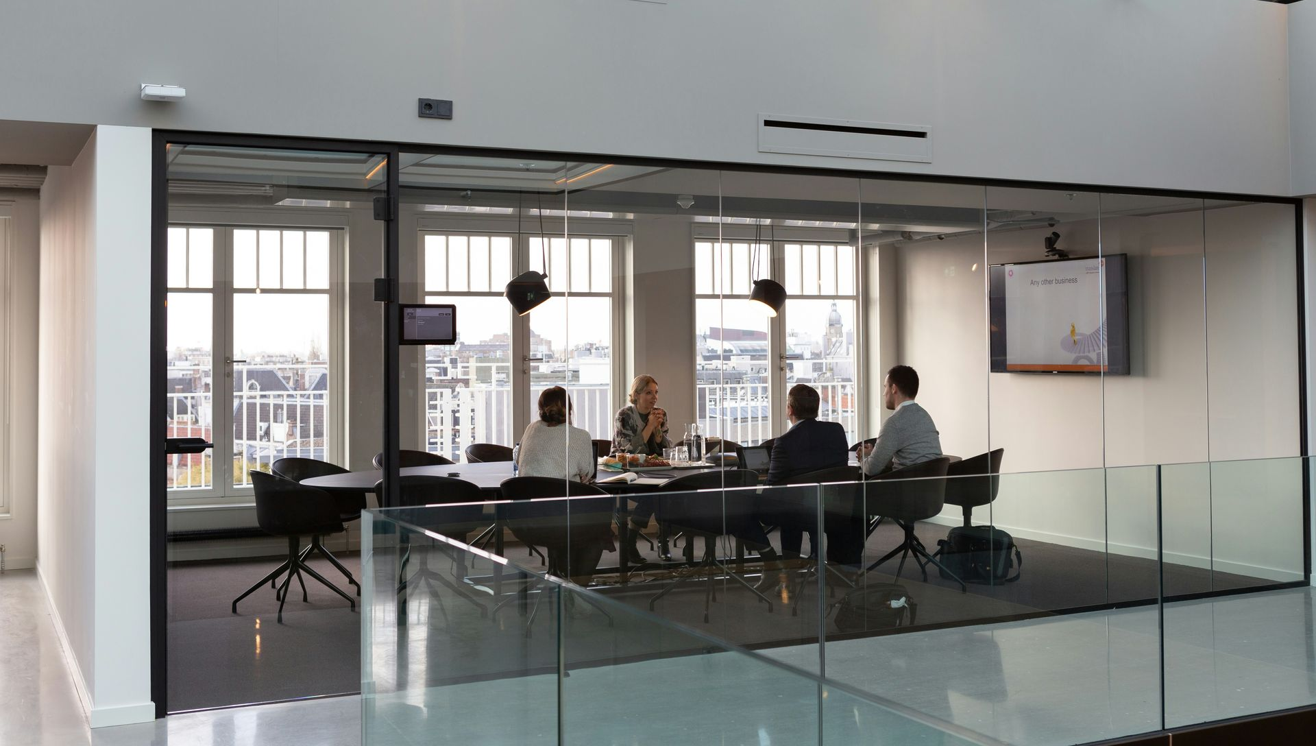 A modern glass-walled conference room with people seated at a table. Sunlight streams in from windows.