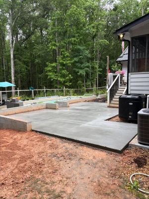 Concrete patio beside a house, with built-in stone seating and wooded backyard behind it.