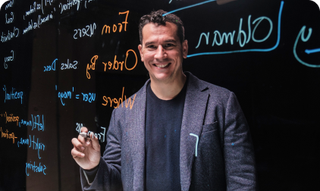 Man smiling, pointing at a whiteboard with colorful handwritten notes.