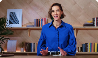 Woman in blue shirt speaking, gesturing with hands; shelves with books and art in the background.