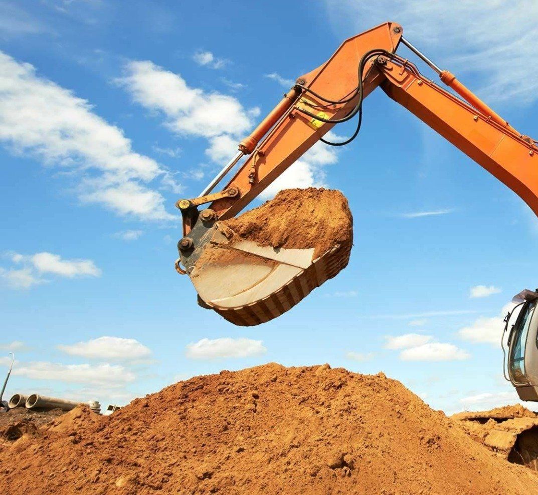 An orange excavator lifts a scoop of brown soil against a blue sky with white clouds, working on a dirt pile.