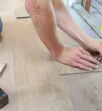 Person installing wood flooring, using a pencil to mark a plank. A hammer sits nearby.