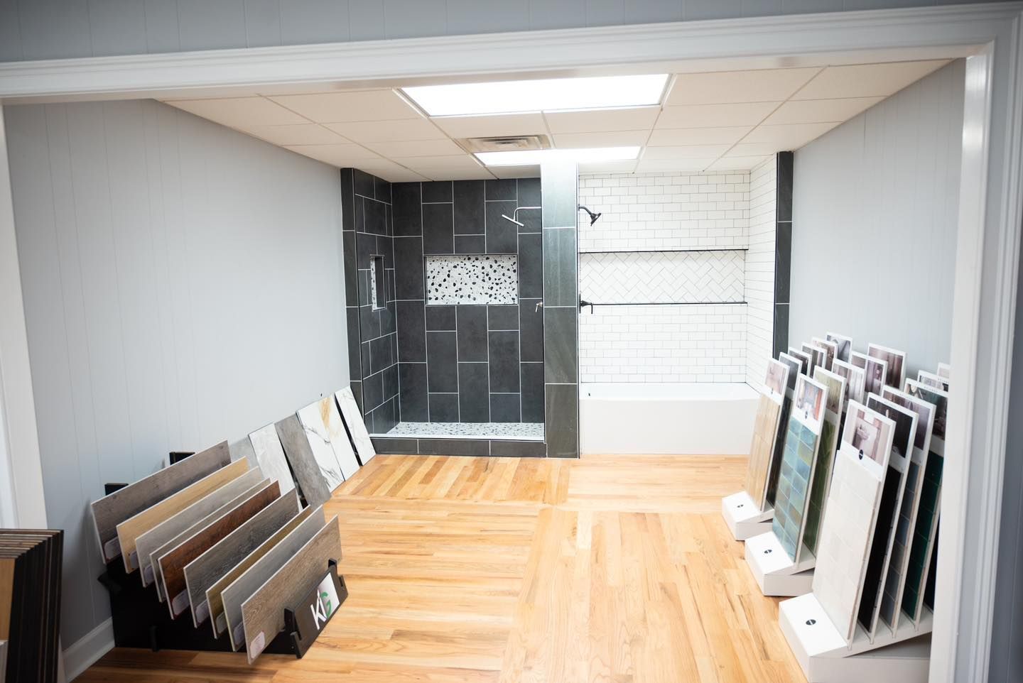 Tile showroom with sample displays and a shower exhibit, wood floor, and gray walls.