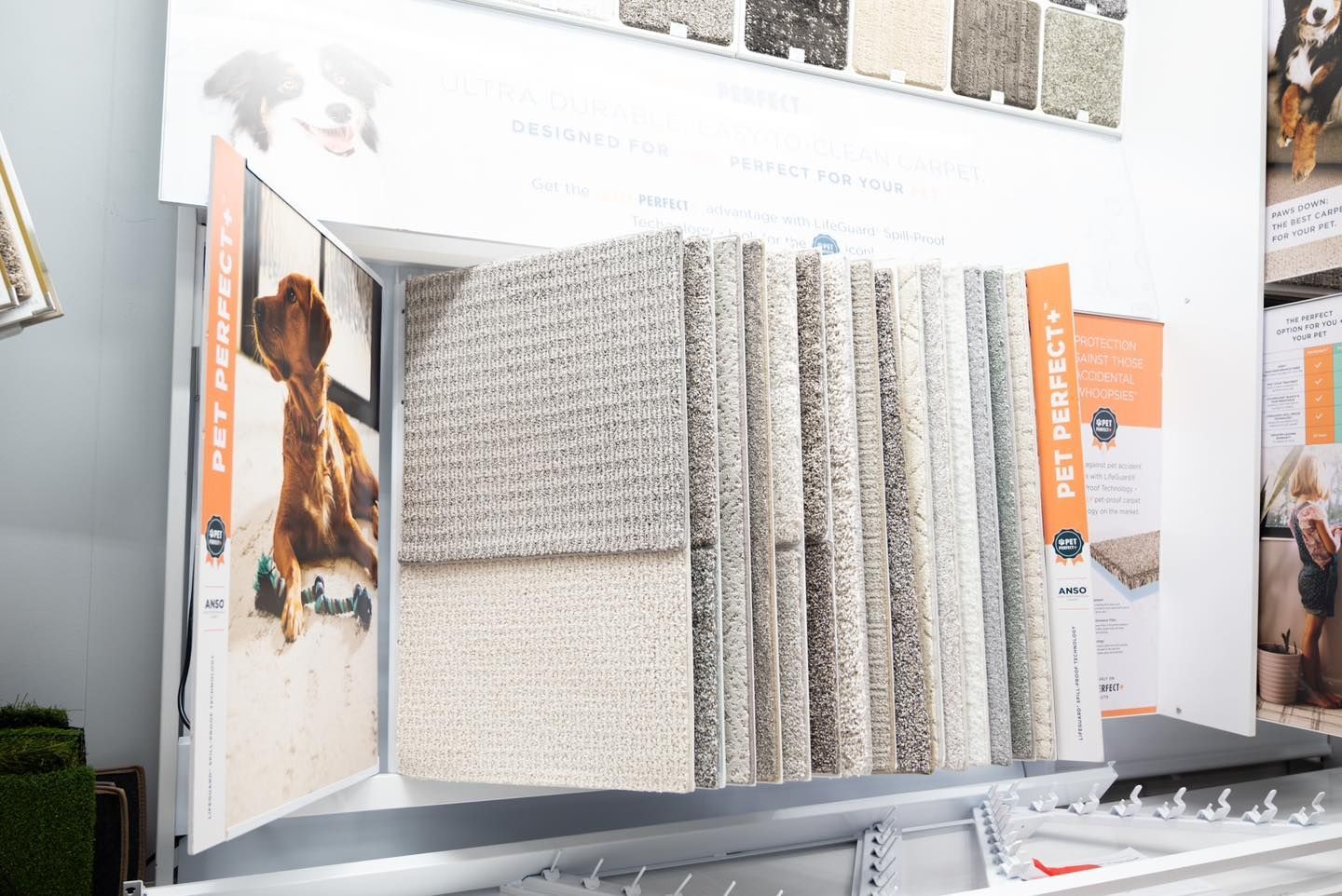 Carpet samples displayed in a store, light neutrals. Signage for pet-friendly options.