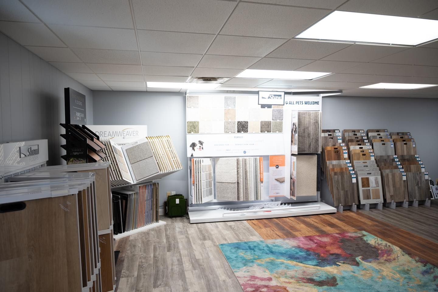 Flooring showroom with wood, tile, and carpet samples. Displays against gray walls and light-colored floor.