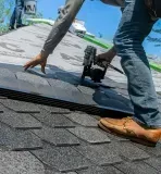 Roofer in jeans and work boots using a nail gun to install asphalt shingles on a roof under a blue sky.
