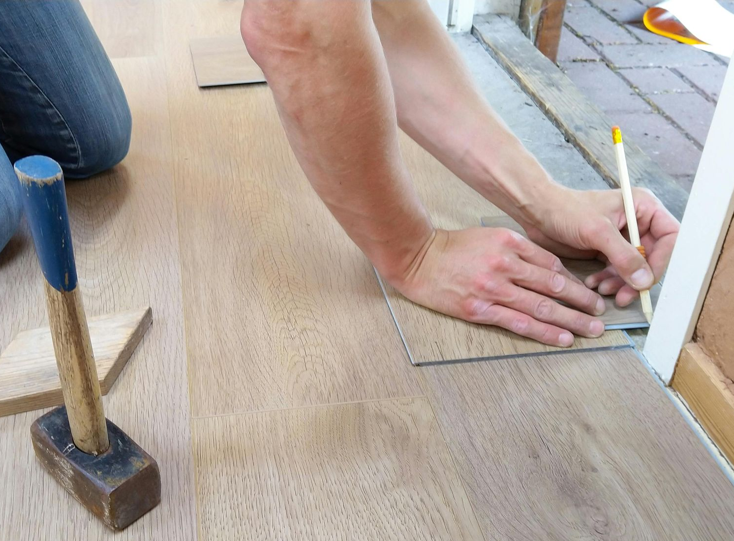 Person installing wood flooring, using a pencil to mark a plank. A hammer sits nearby.