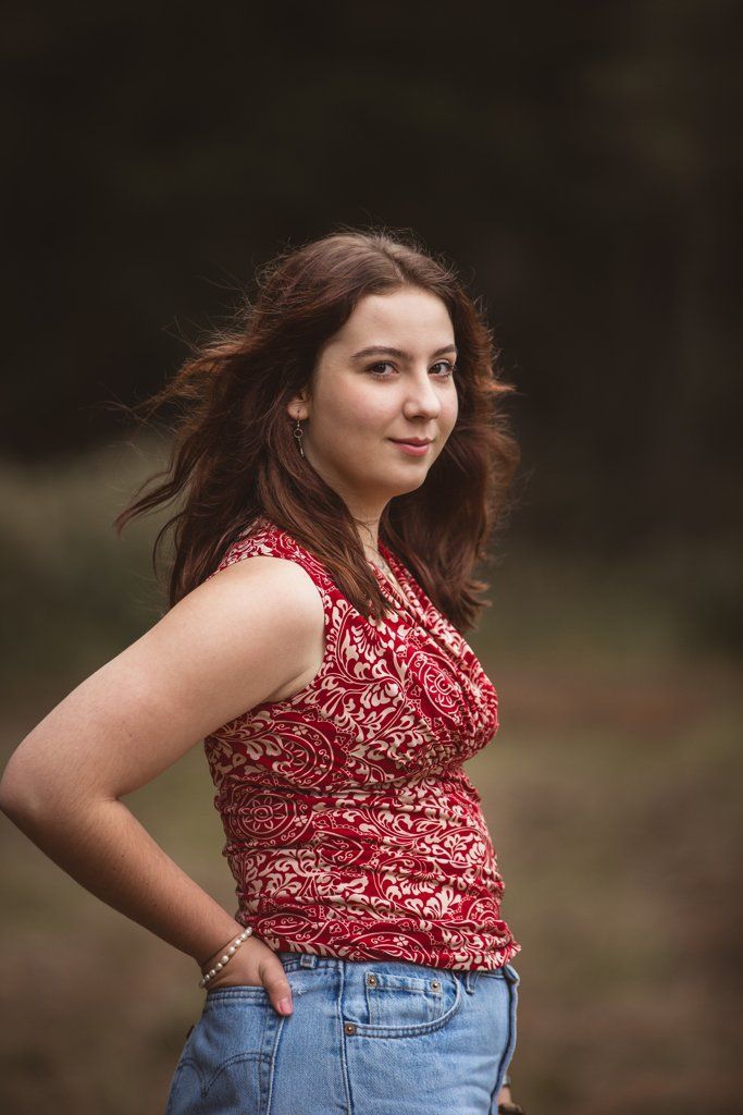 A young woman in a red tank top and blue jeans is standing in a field.