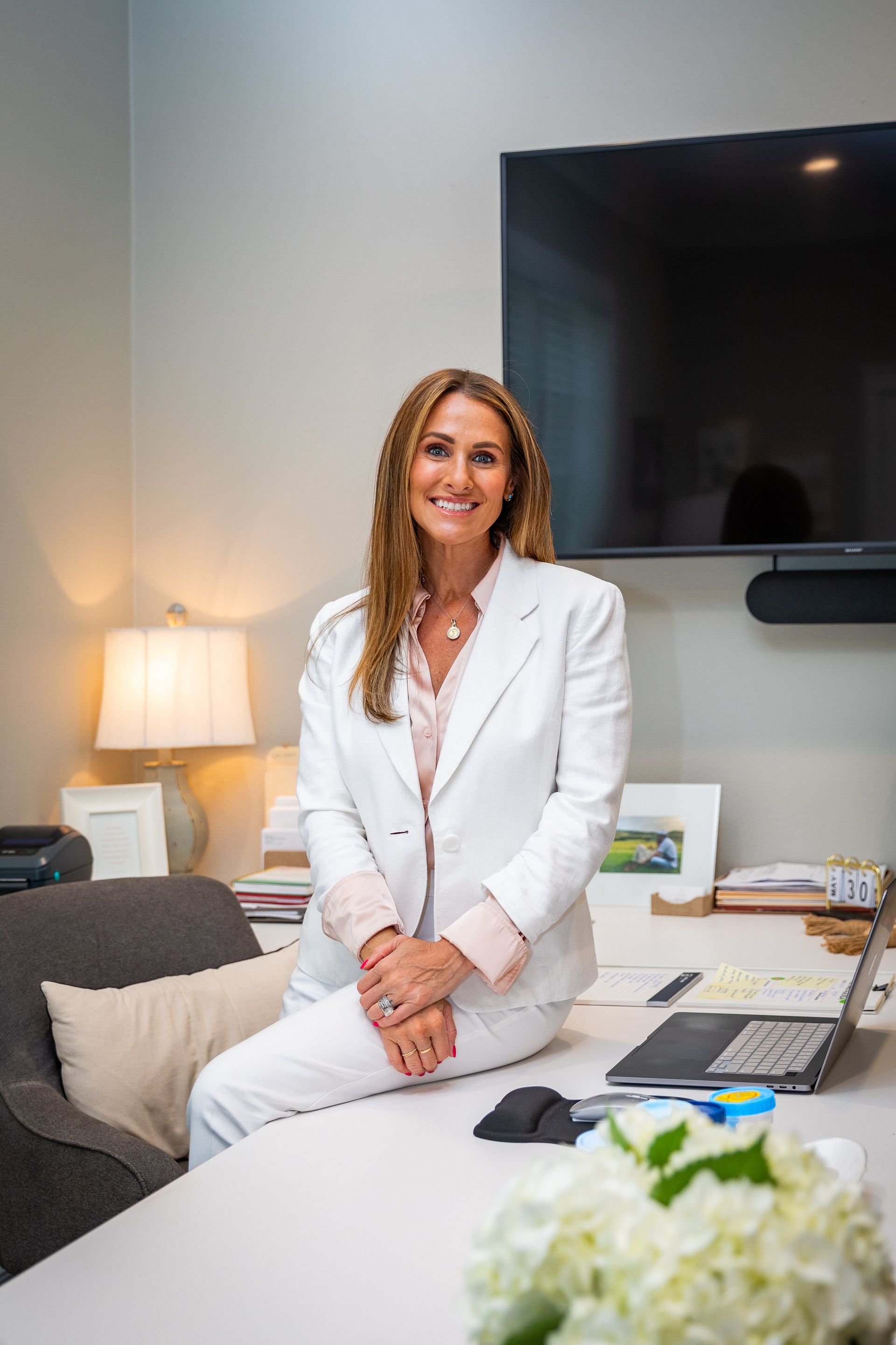 A woman in a white suit is sitting at a desk in front of a television.