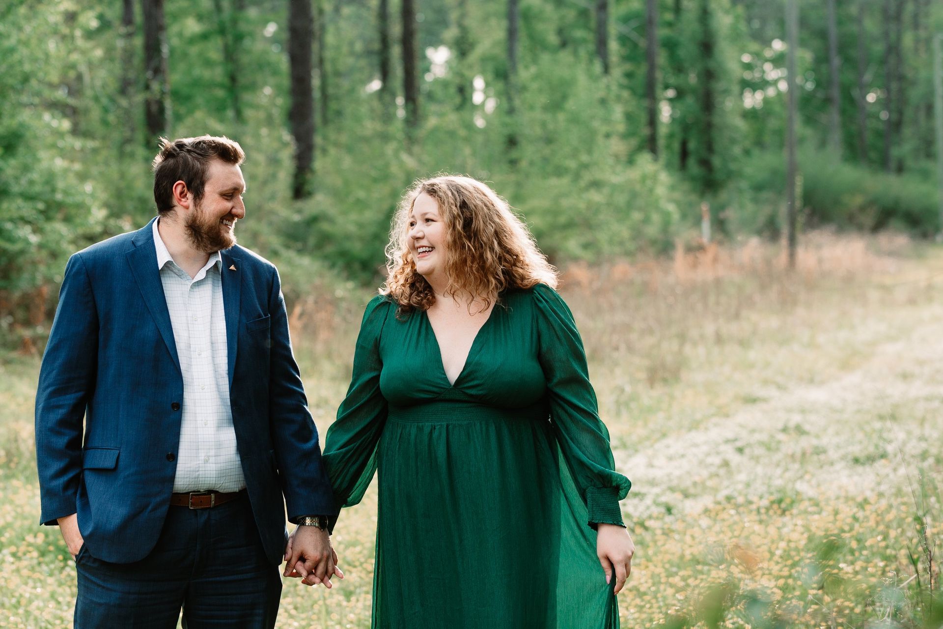 A man in a suit and a woman in a green dress are holding hands in the woods.