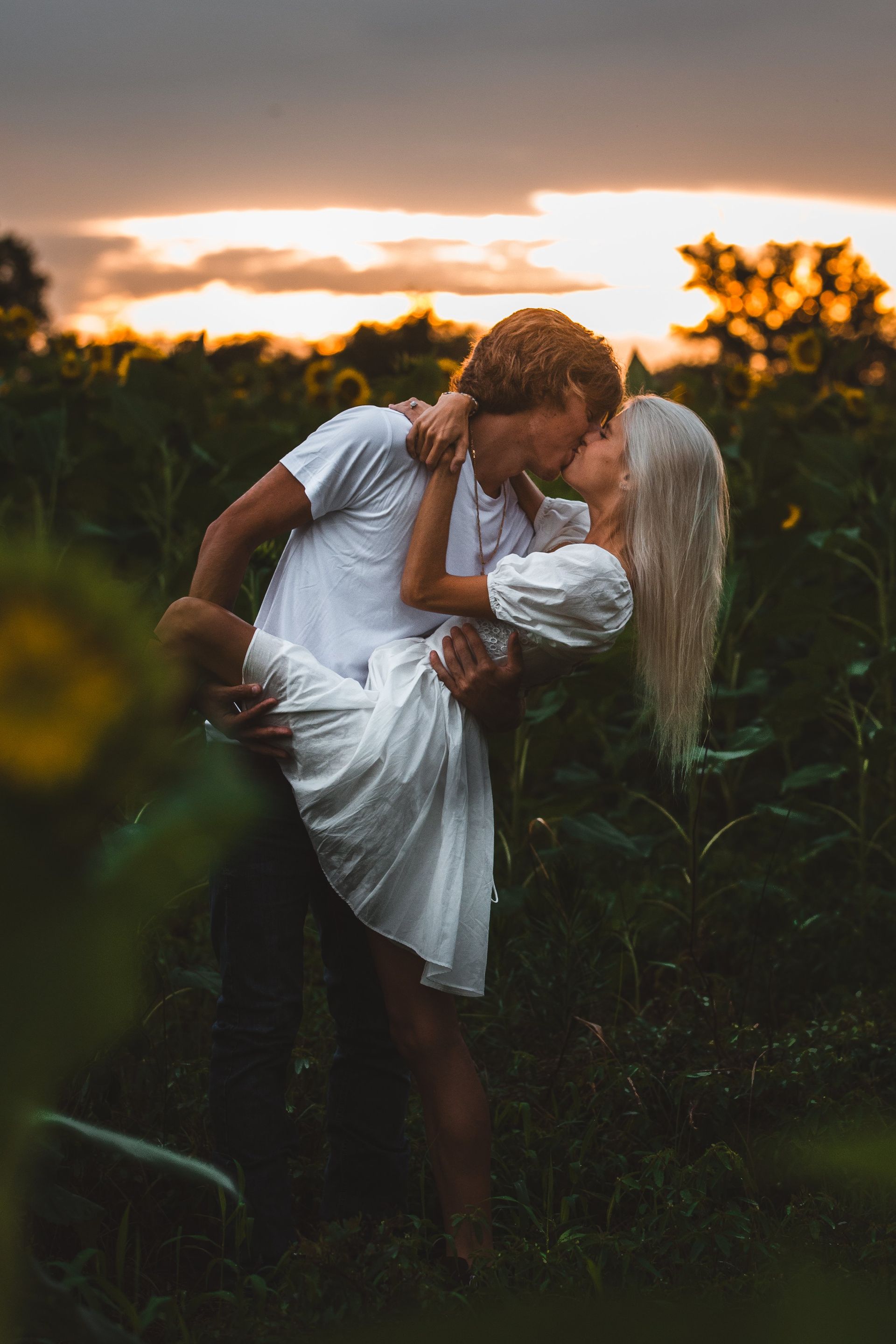 A man is holding a woman in his arms and kissing her in a field of sunflowers.