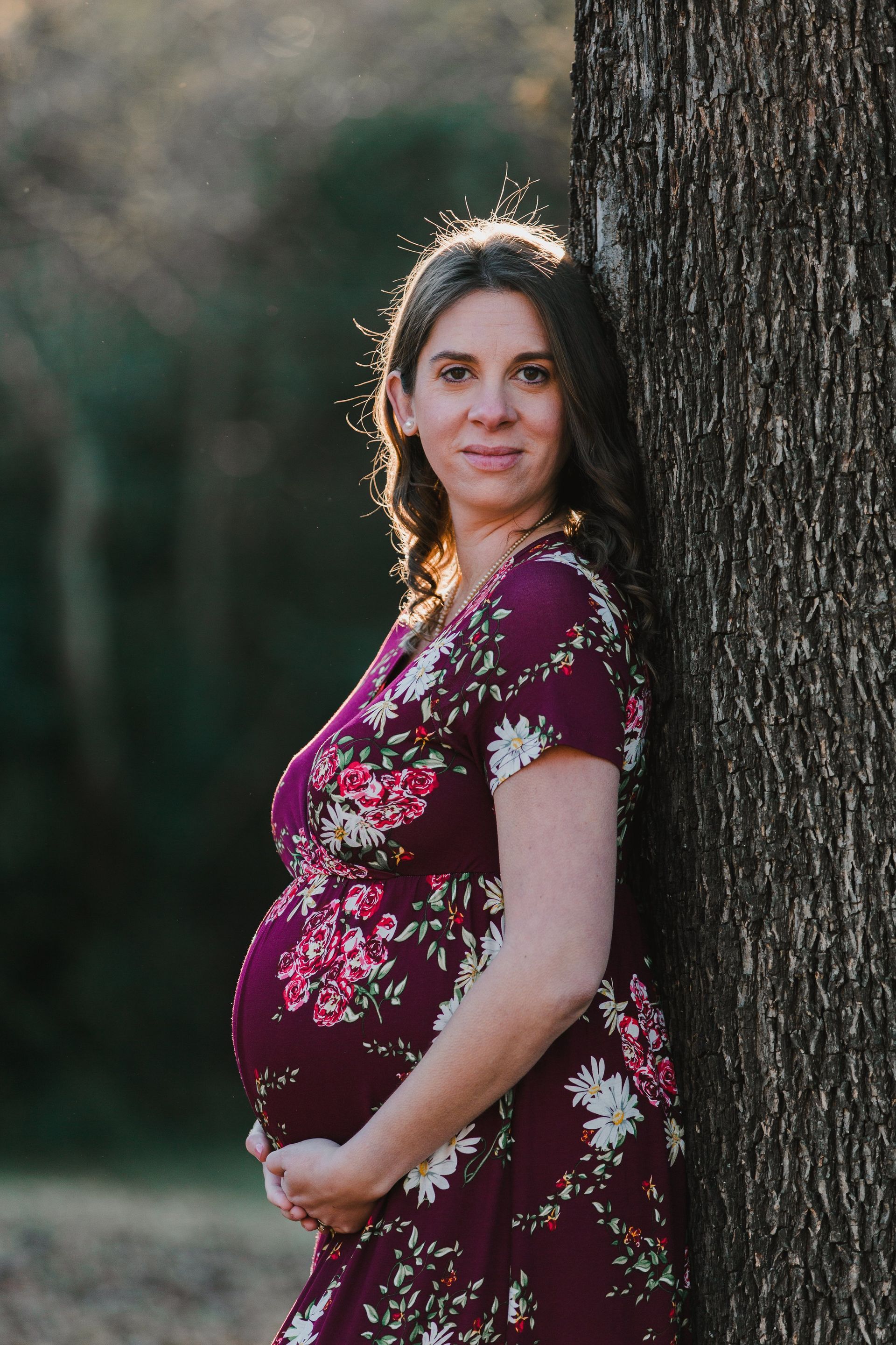 A pregnant woman in a purple dress is leaning against a tree.