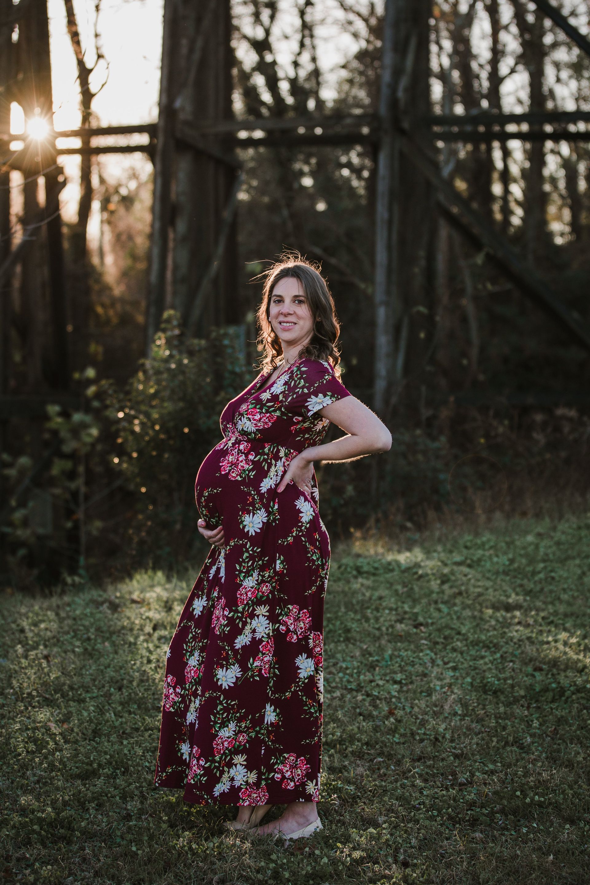A pregnant woman in a floral dress is standing in the grass.