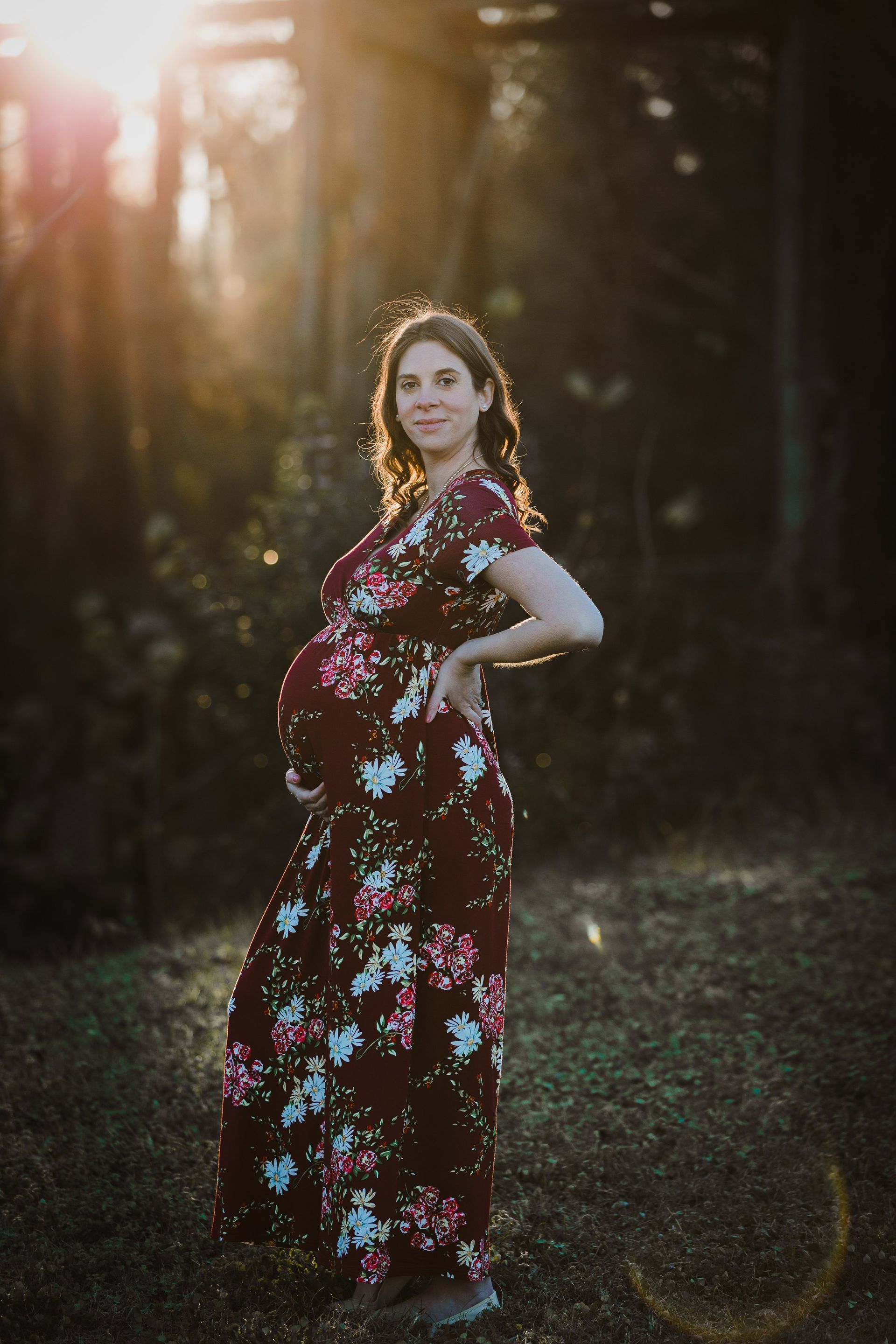 A pregnant woman in a floral dress is standing in the woods with her hands on her hips.
