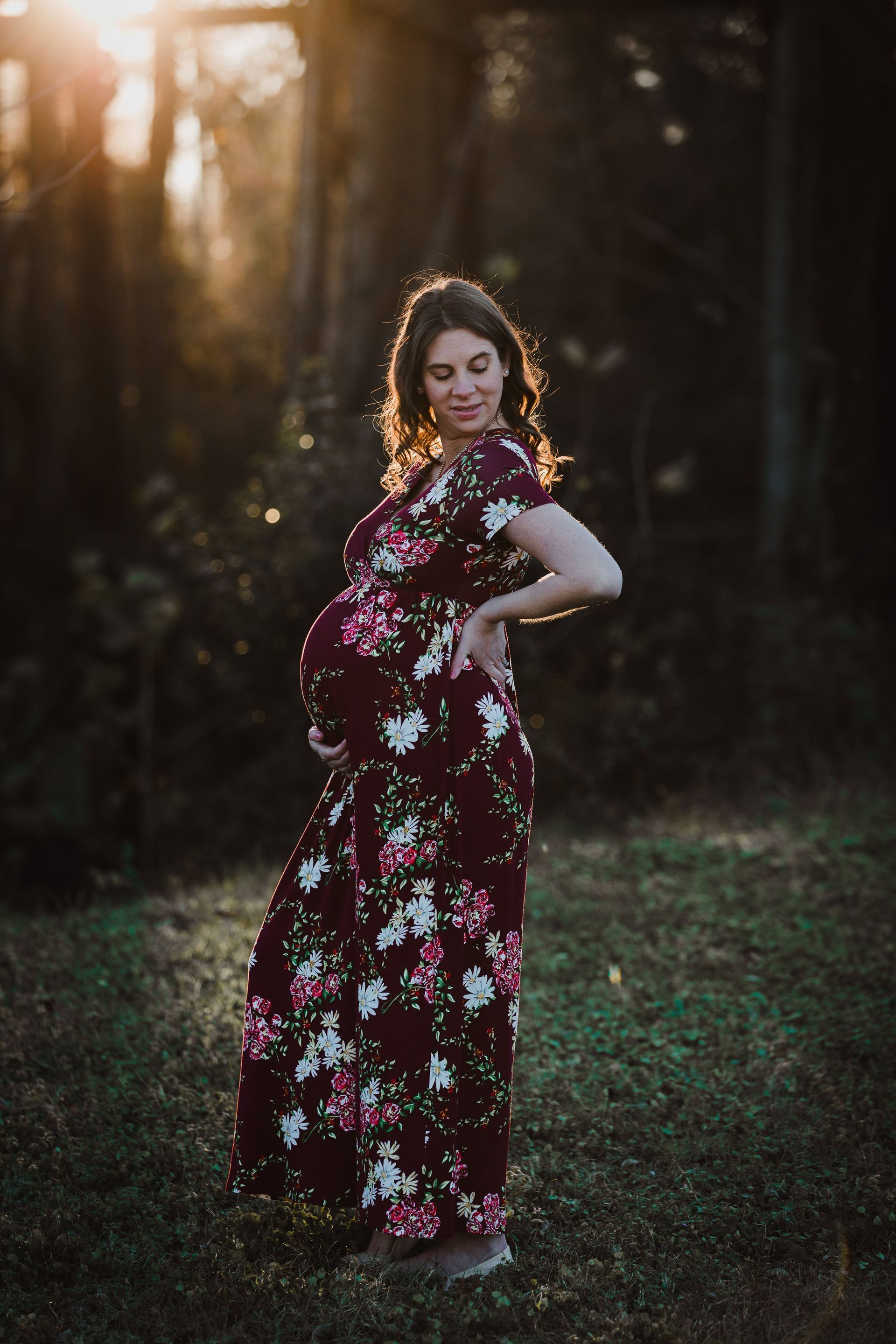 A pregnant woman in a floral dress is standing in the grass holding her belly.