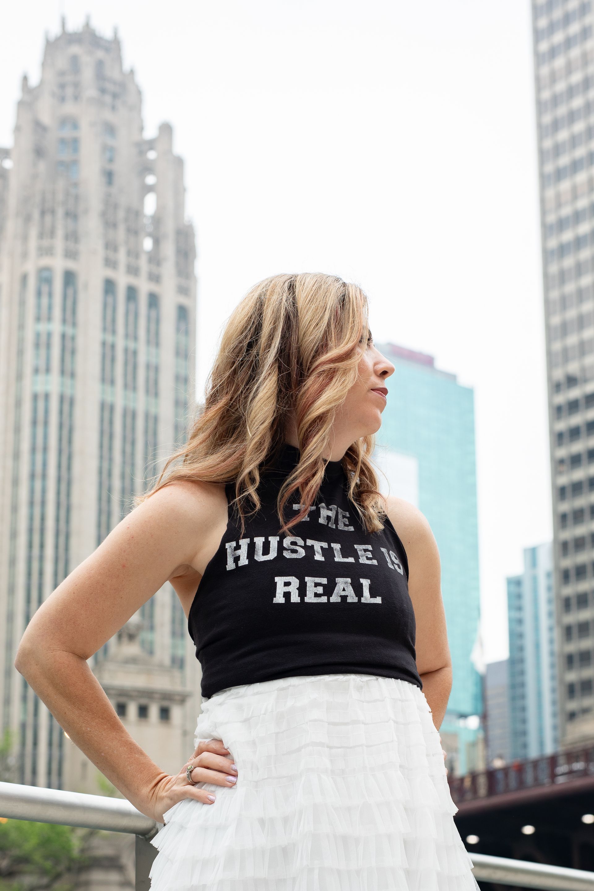 A woman wearing a black tank top that says hustle 's real is standing in front of a city skyline.