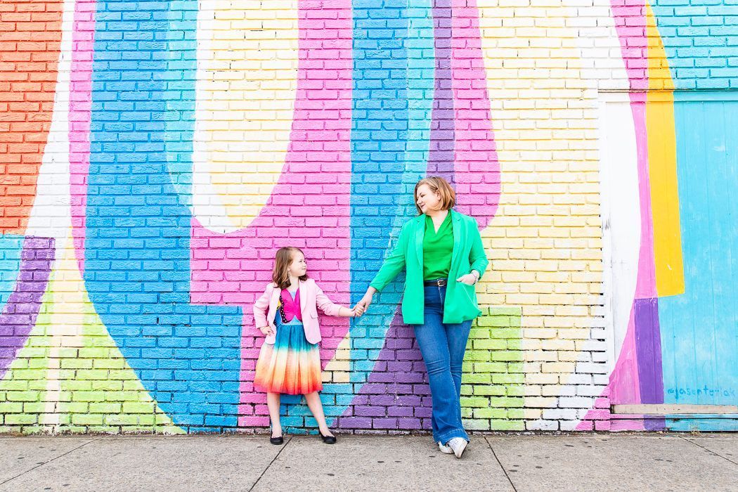 A woman and a little girl are standing in front of a colorful brick wall holding hands.
