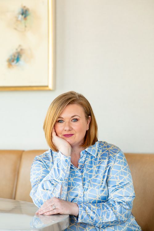 A woman in a blue shirt is sitting at a table with her hand on her chin.