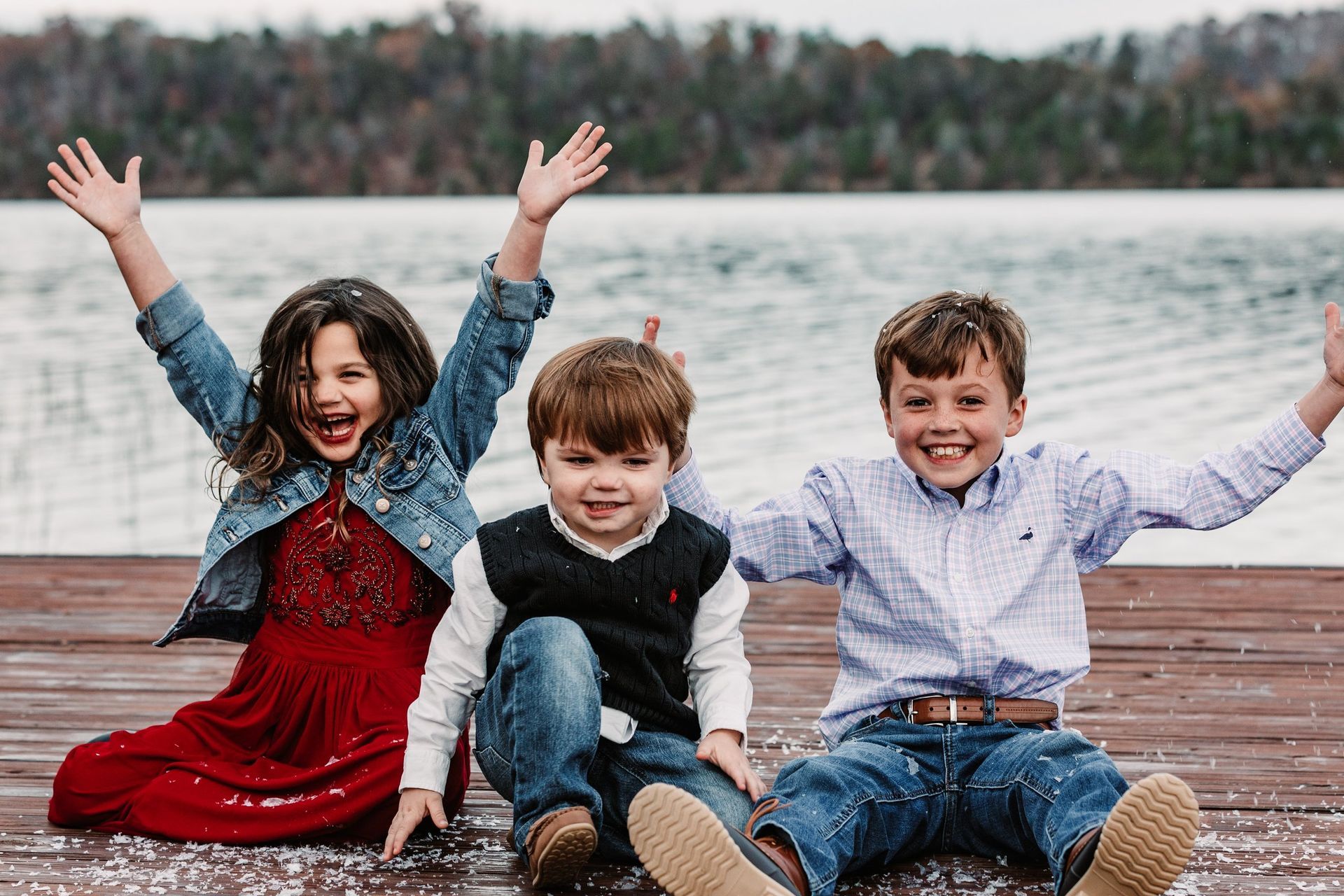 Three children are sitting on a dock with their arms in the air.