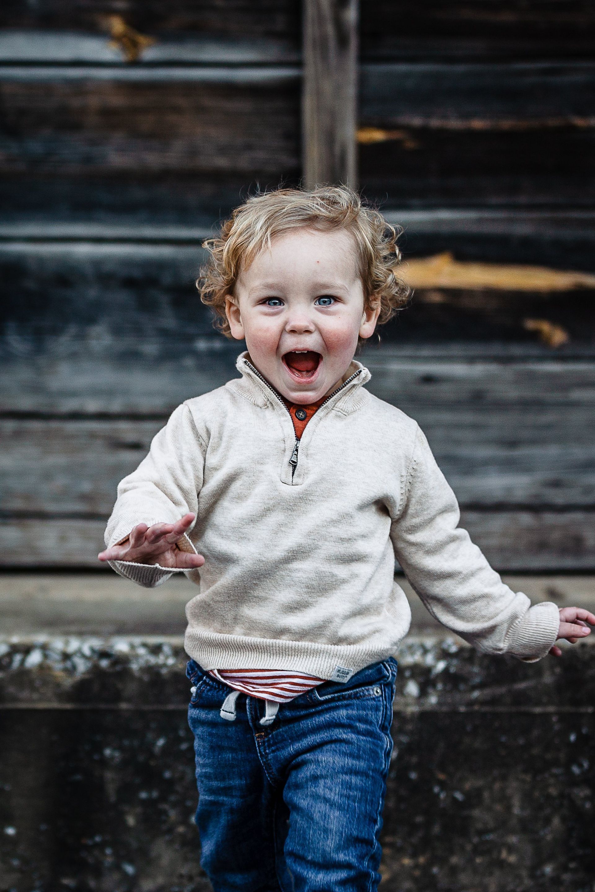 A little boy with curly hair is standing in front of a wooden building.