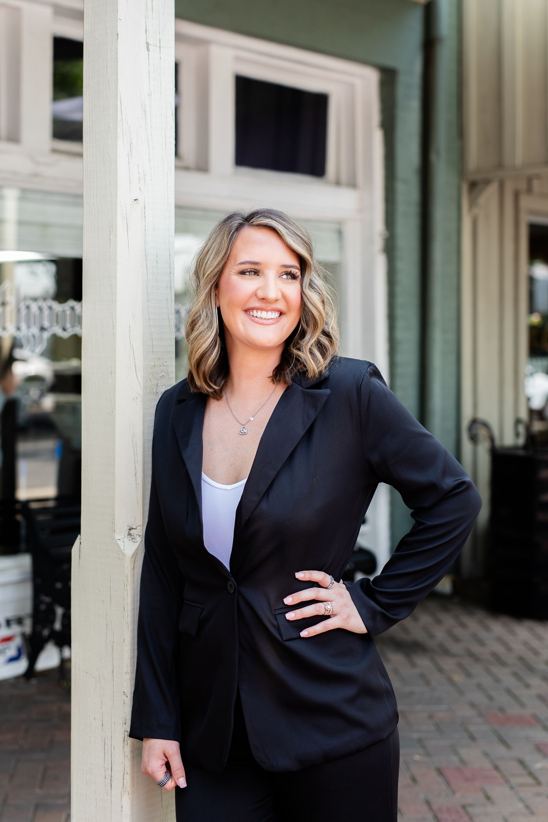 A woman in a black suit is leaning against a pole.