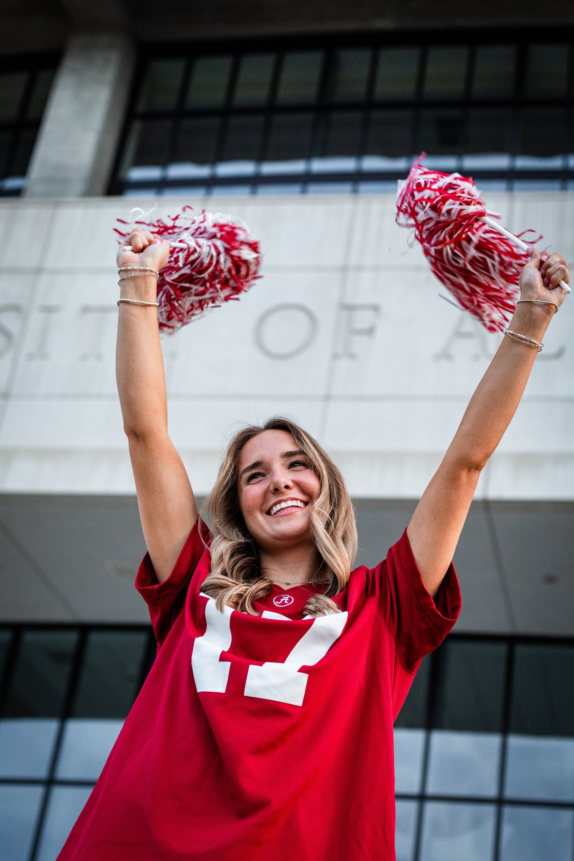 A woman in a red shirt is holding pom poms in her hands in front of a building.